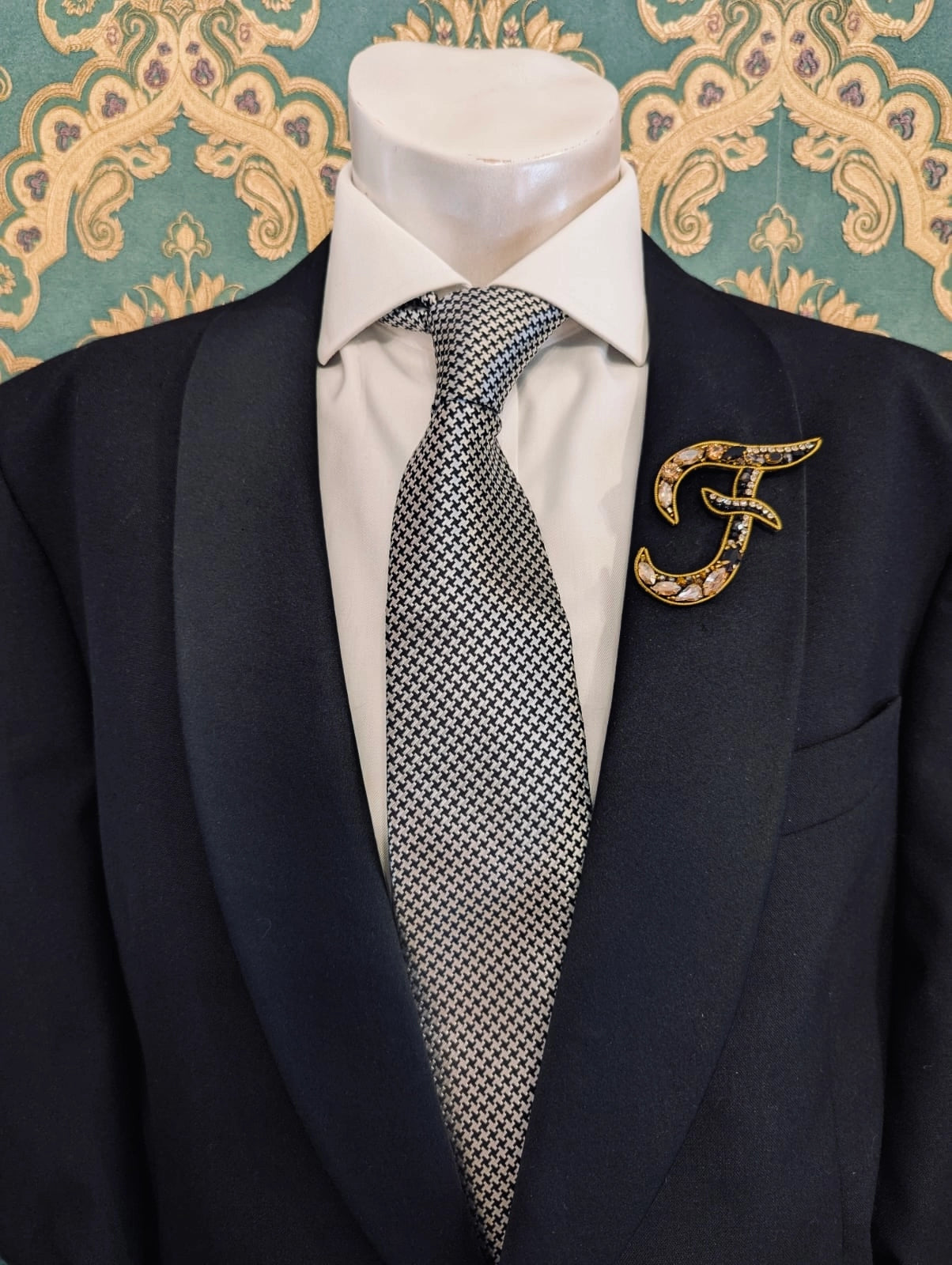 Broochella men's hand beaded F initial brooch in black and gold beads and sequins. Pinned to a men's mannequin wearing a black suit and a tie. The background is green and gold damask wallpaper.