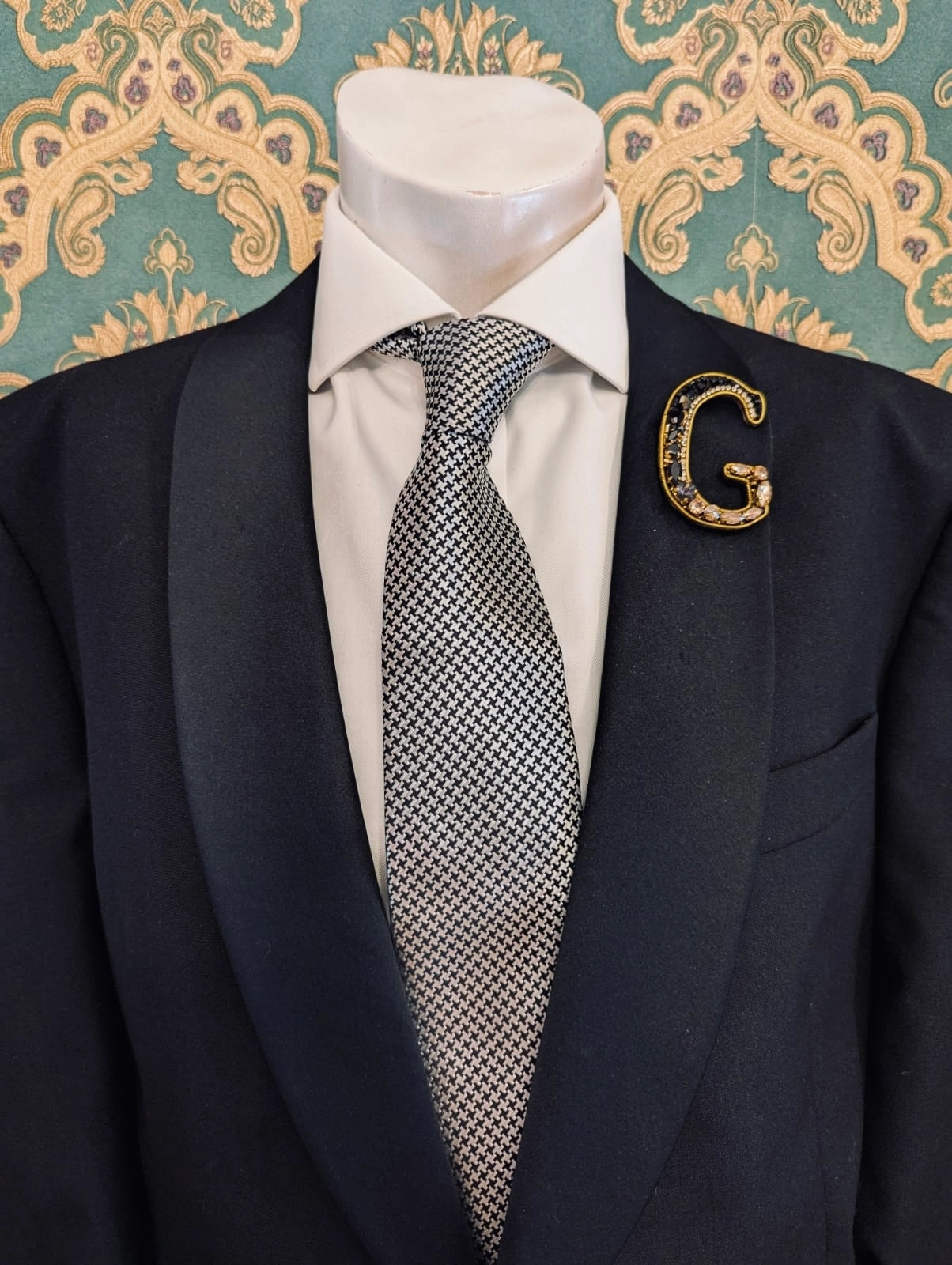 Broochella men's hand beaded G initial brooch in black and gold beads and sequins. Pinned to a men's mannequin wearing a black suit and a tie. The background is green and gold damask wallpaper.