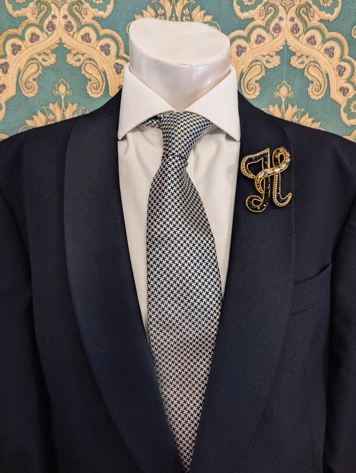 Broochella men's hand beaded H initial brooch in black and gold beads and sequins. Pinned to a men's mannequin wearing a black suit and a tie. The background is green and gold damask wallpaper.