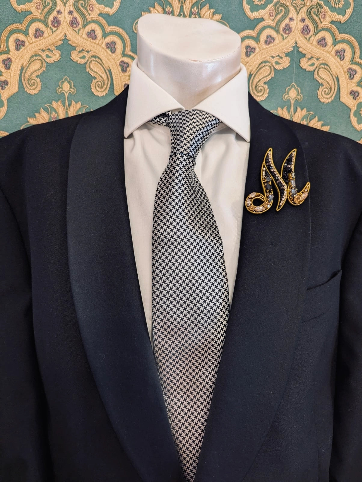 Broochella men's hand beaded M initial brooch in black and gold beads and sequins. Pinned to a men's mannequin wearing a black suit and a tie. The background is green and gold damask wallpaper.