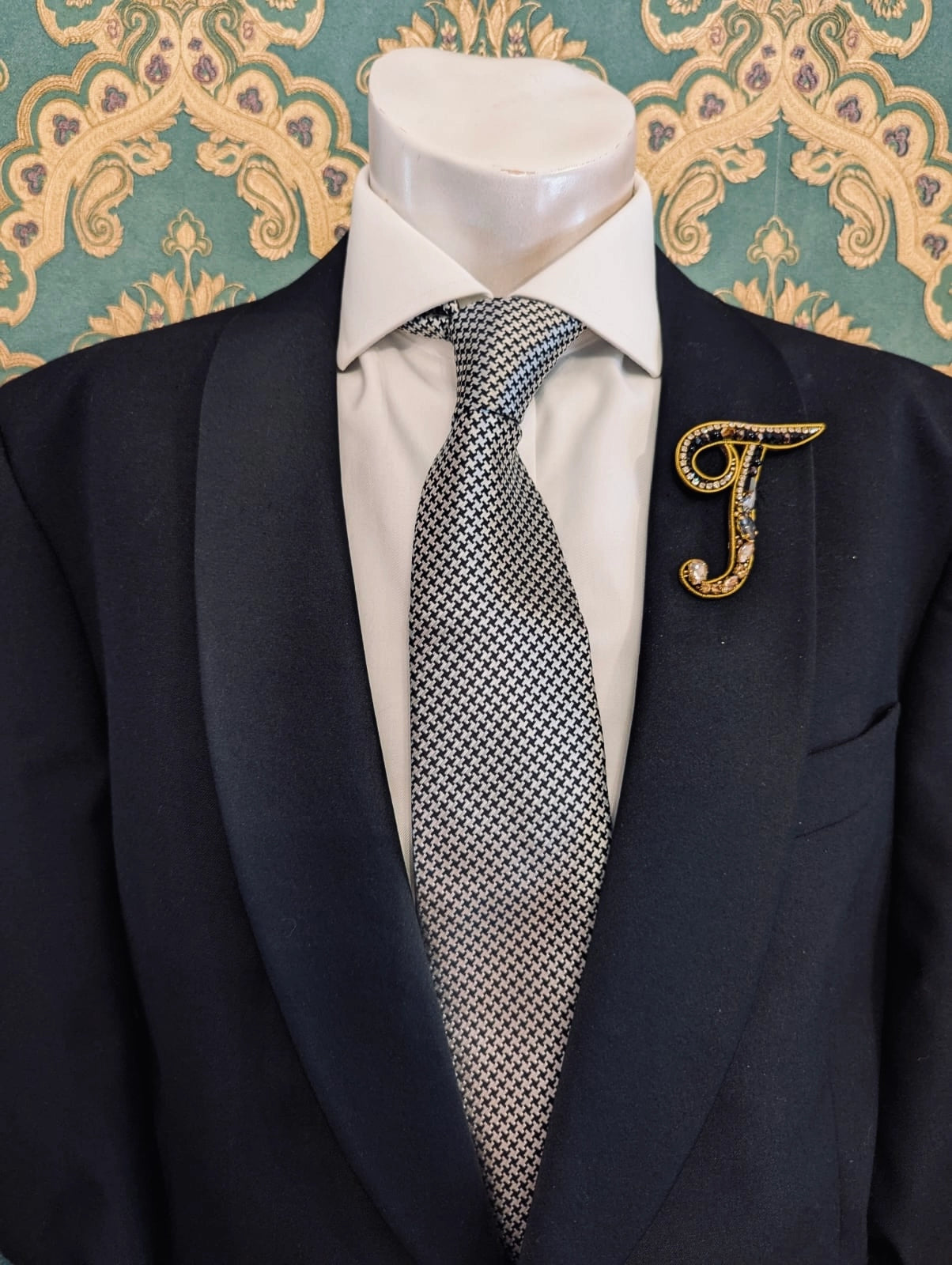 Broochella men's hand beaded T initial brooch in black and gold beads and sequins. Pinned to a men's mannequin wearing a black suit and a tie. The background is green and gold damask wallpaper.