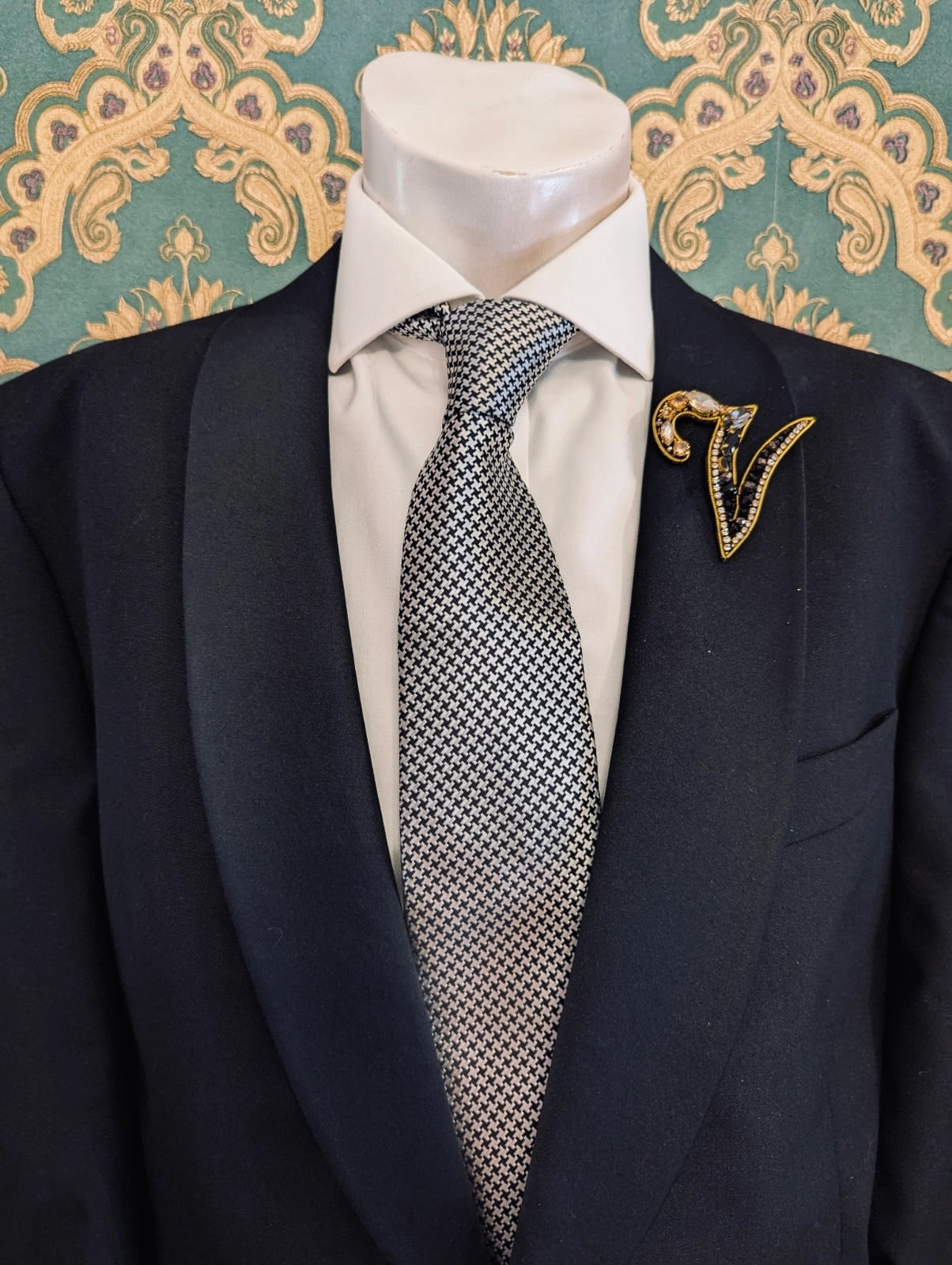 Broochella men's hand beaded B initial brooch in black and gold beads and sequins. Pinned to a men's mannequin wearing a black suit and a tie. The background is green and gold damask wallpaper.
