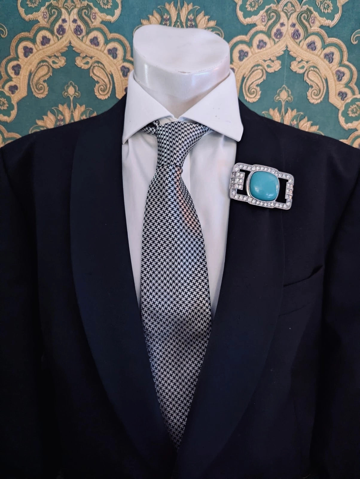 Broochella men's libertene silver metal brooch with crystals and turquoise centre. Pinned to a men's mannequin wearing a black suit and houndstooth tie. The background is green and gold damask wallpaper.