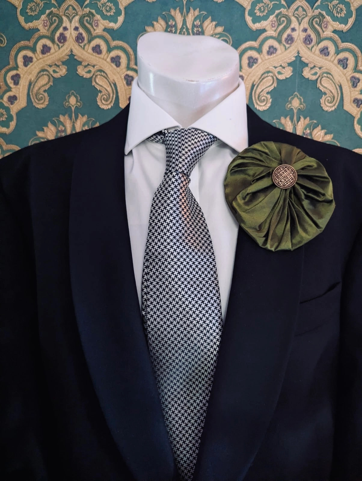 Broochella men's large green handmade silk lollipop brooch with a gold button centre. Pinned to a men's mannequin wearing a black suit and a tie. The background is green and gold damask wallpaper.