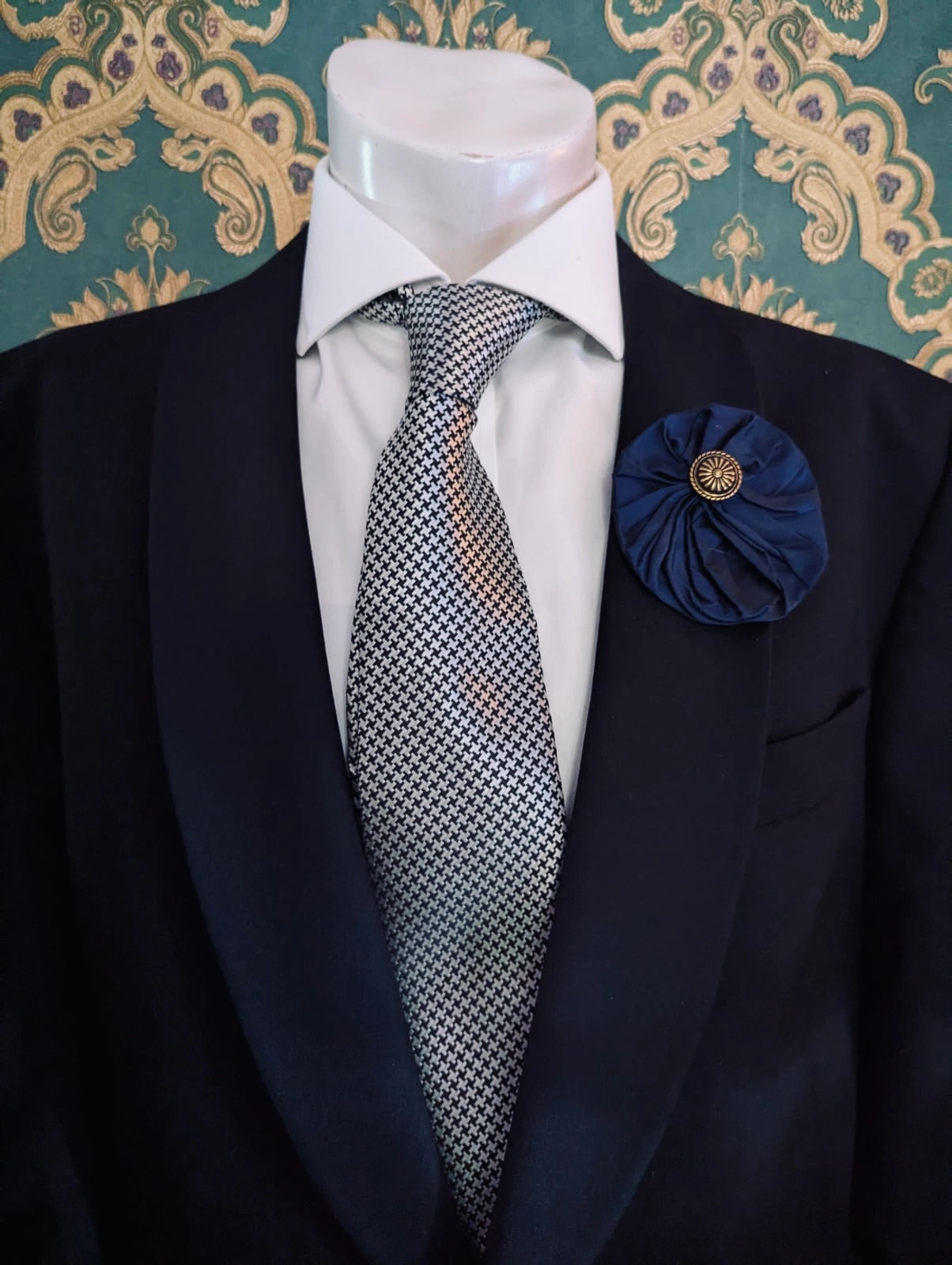 Broochella men's medium navy handmade silk lollipop brooch with a gold button centre. Pinned to a men's mannequin wearing a black suit and a tie. The background is green and gold damask wallpaper.