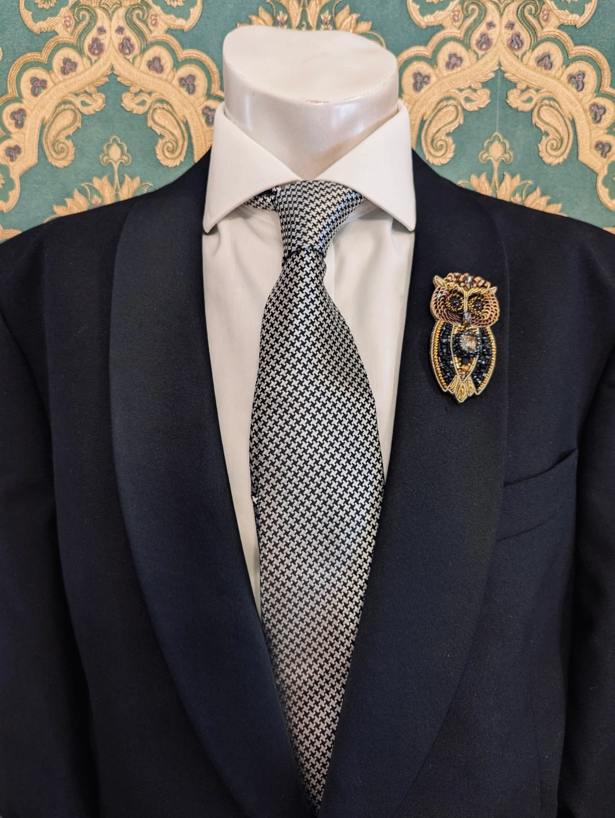 Broochella men's hand beaded owl brooch in black and gold. Pinned to a men's mannequin wearing a black suit and a tie. The background is green and gold damask wallpaper.