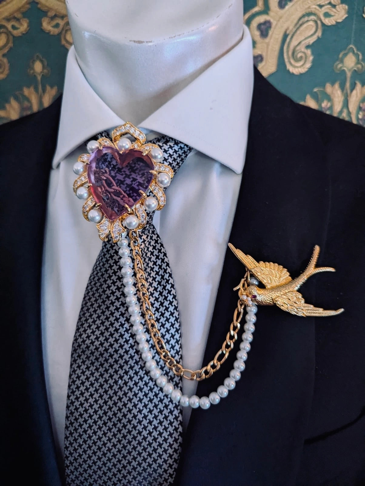 Broochella men's romeo and juliet heart shaped lilac crystal brooch, with gold metal and pearl frame. linked to the broochella swallow brooch with a pearl and gold chain. Pinned to a men's mannequin wearing a black suit and houndstooth tie. The background is green and gold damask wallpaper.