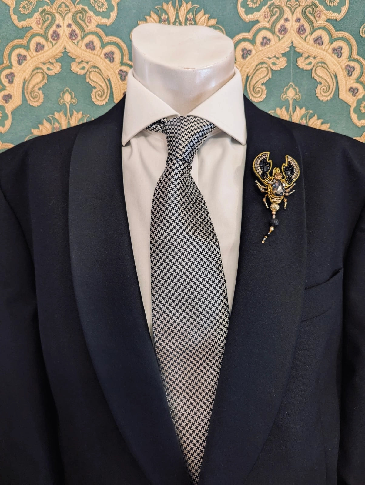 Broochella men's hand beaded scorpion brooch in black and gold. Pinned to a men's mannequin wearing a black suit and a tie. The background is green and gold damask wallpaper.