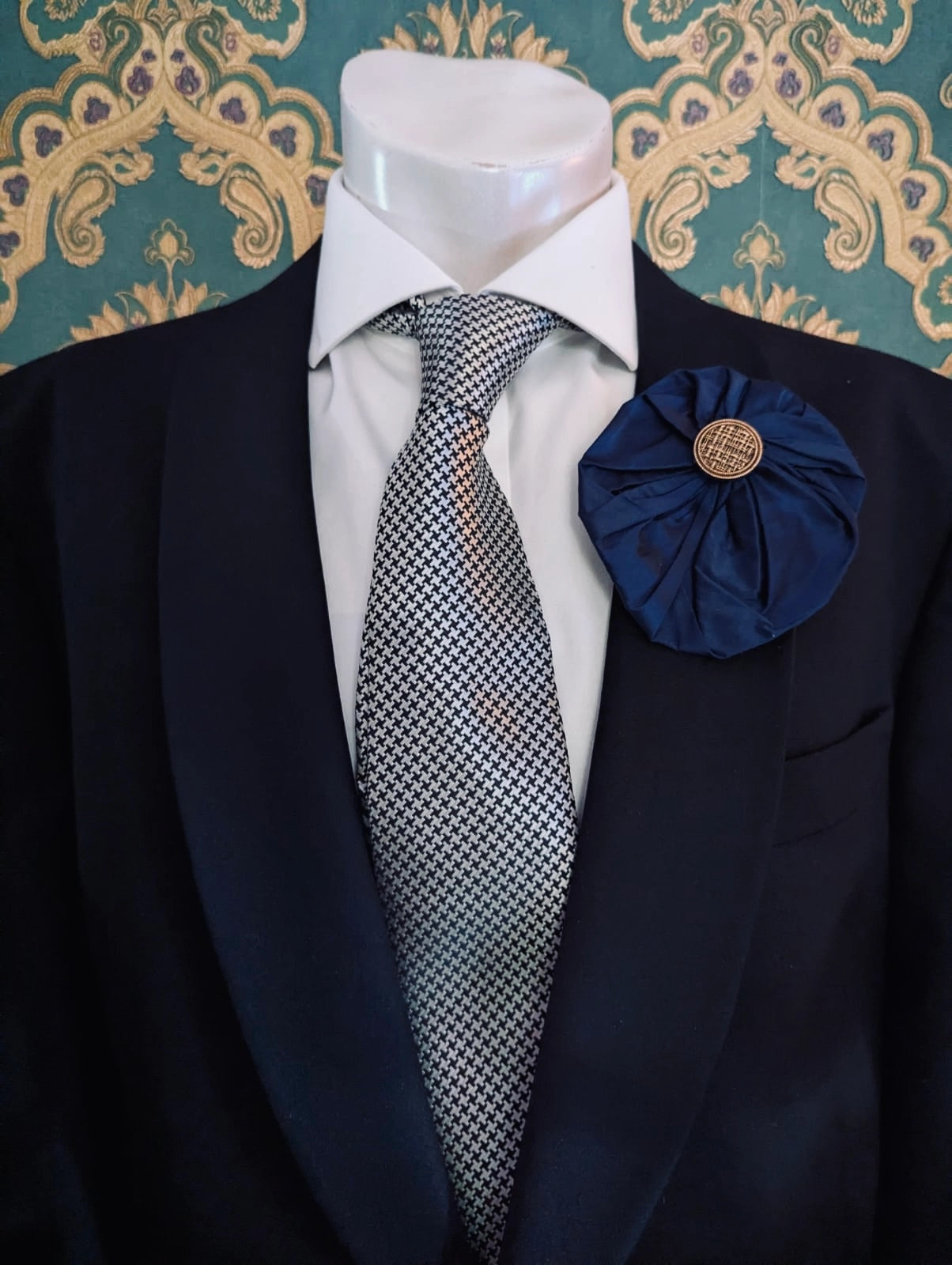 Broochella men's large navy handmade silk lollipop brooch with a gold button centre. Pinned to a men's mannequin wearing a black suit and a tie. The background is green and gold damask wallpaper.