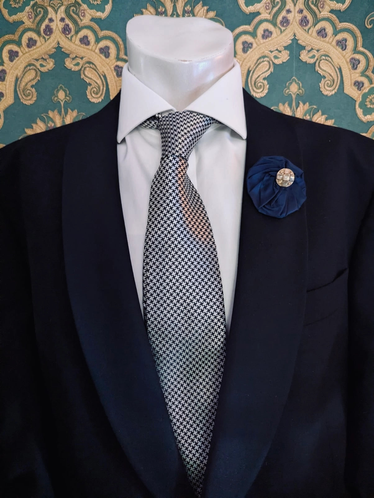 Broochella men's small navy handmade silk lollipop brooch with a gold button centre. Pinned to a men's mannequin wearing a black suit and a tie. The background is green and gold damask wallpaper.