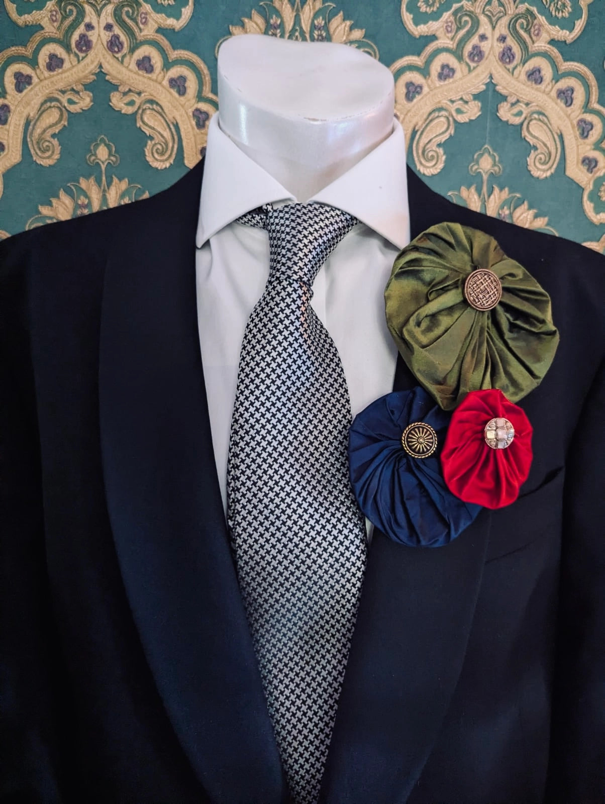 Broochella  men's silk lollipop brooches in red, navy and green. They are pinned to a men's mannequin wearing a black suit and a tie. The background is green and gold damask wallpaper.