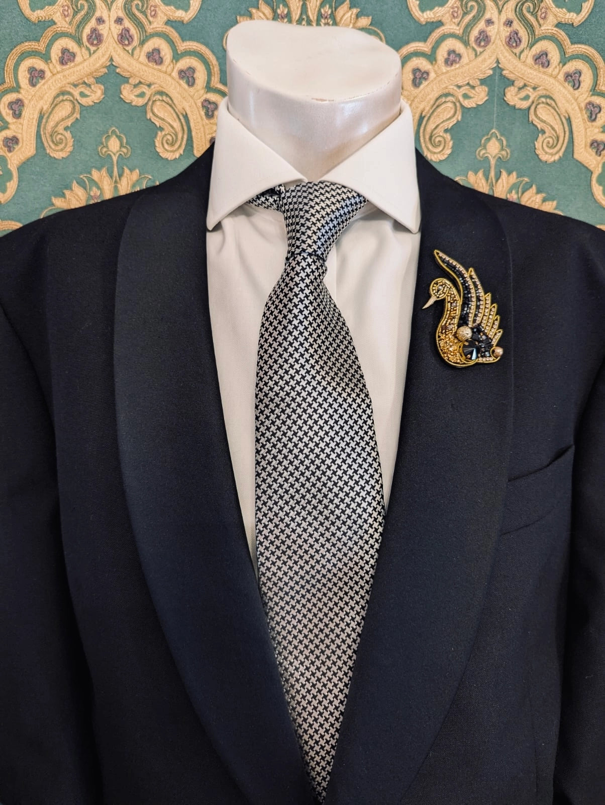 Broochella men's hand beaded swan brooch in black and gold. Pinned to a men's mannequin wearing a black suit and a tie. The background is green and gold damask wallpaper.