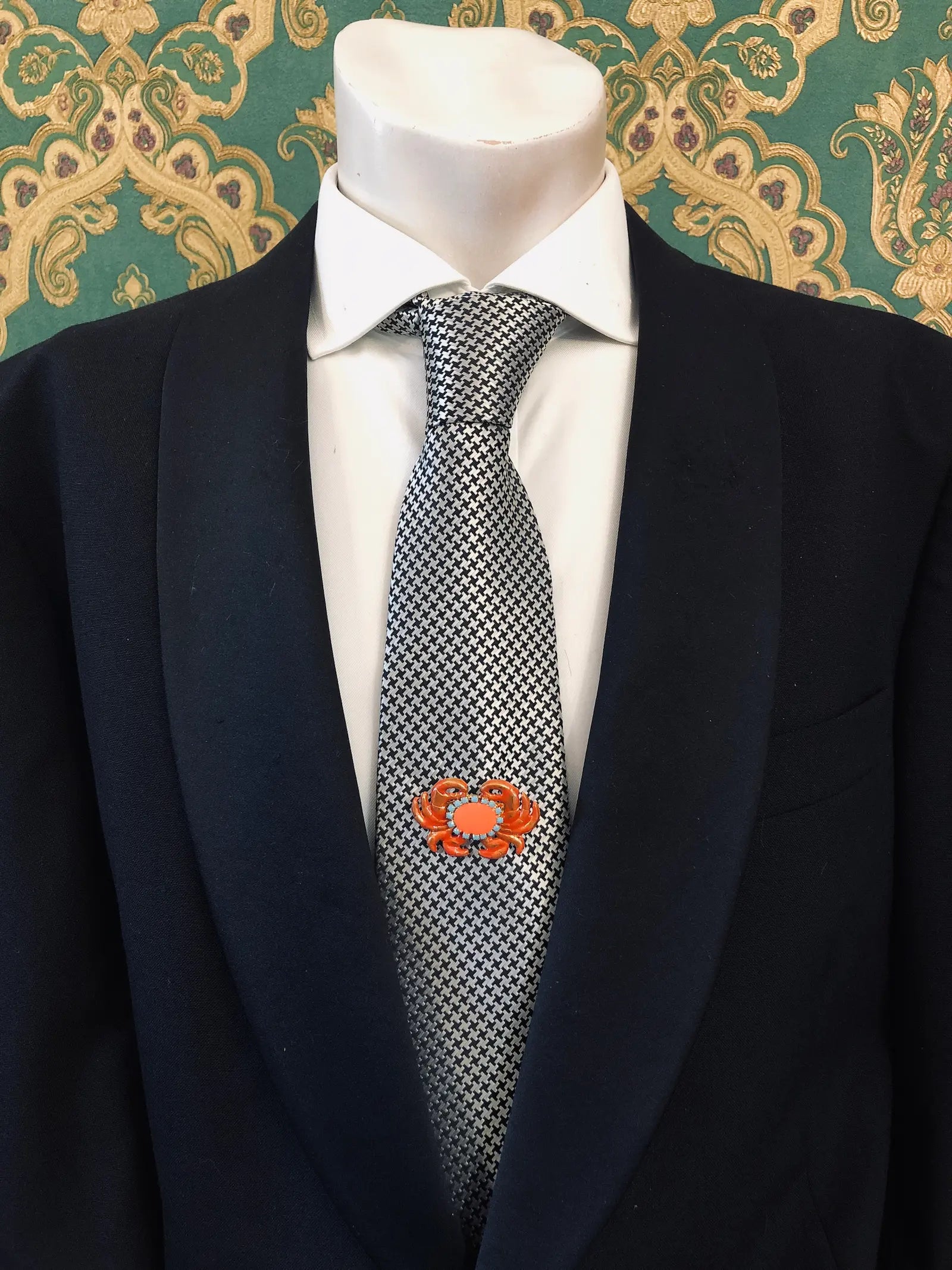 A close-up of a mannequin torso wearing a black suit jacket, white dress shirt, and a black-and-white patterned tie. A Broochella Men's Sacro Cuore  small orange crab-shaped lapel brooch is pinned on the left chest.