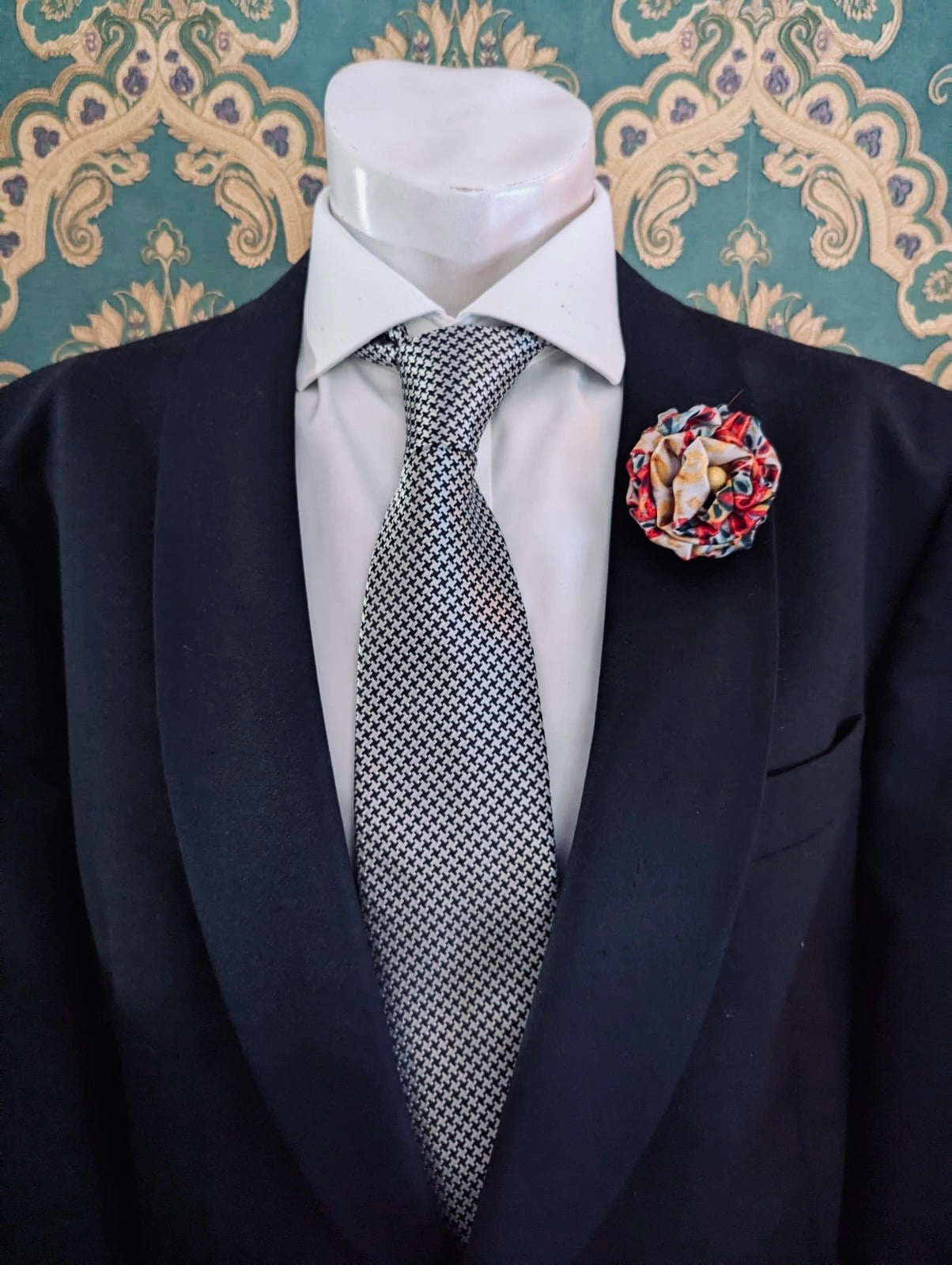 A men's formal outfit displayed on a white mannequin torso, featuring a black suit jacket, a white dress shirt, a black-and-white patterned tie, and a Broochella men's sartorial bloom flower brooch pin against a teal damask-patterned background.