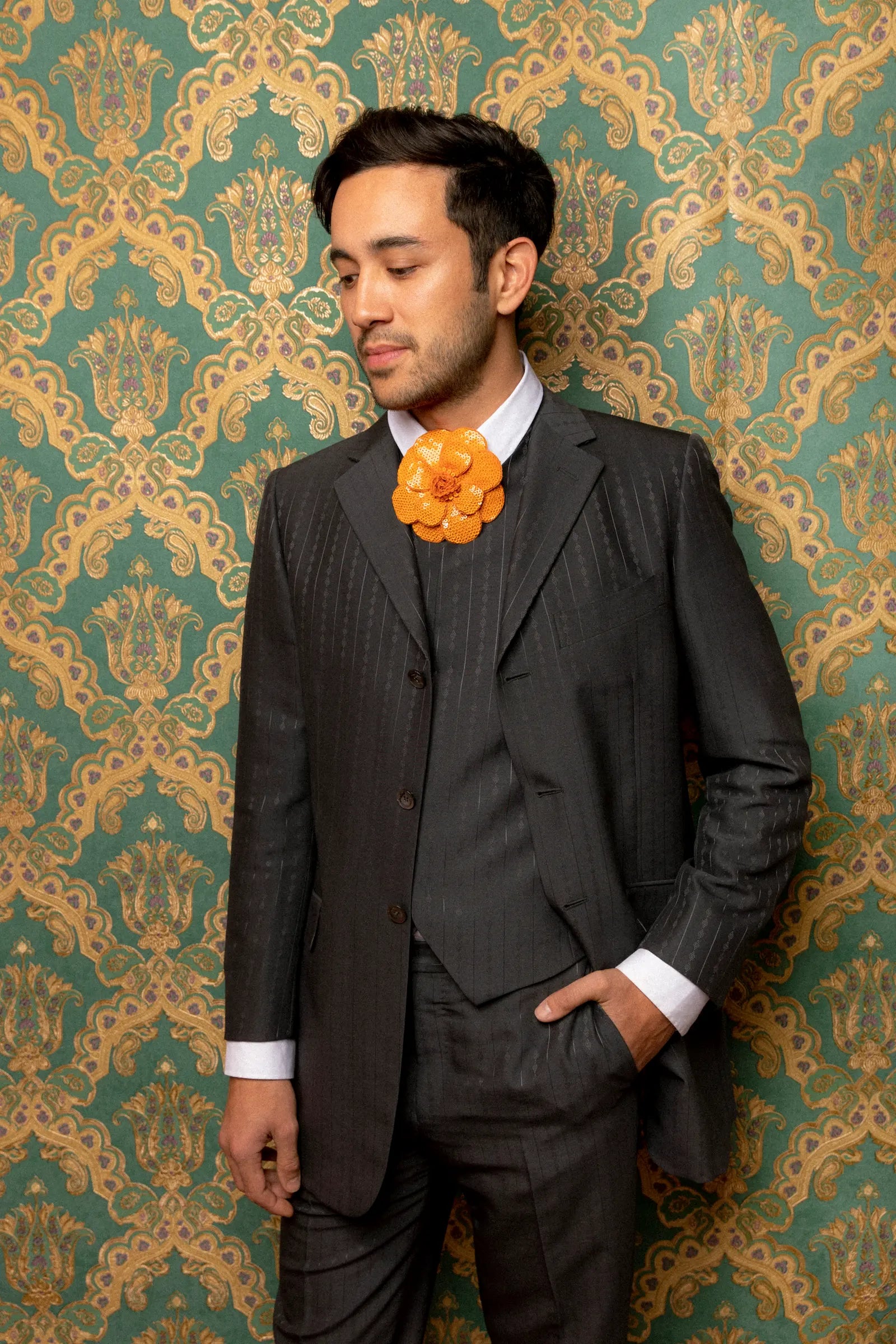 A man wearing a dark gray pinstripe three-piece suit with a white dress shirt and a Broochella Men's Scintillante bright orange flower-shaped necktie, standing against a green and gold damask wallpaper background.