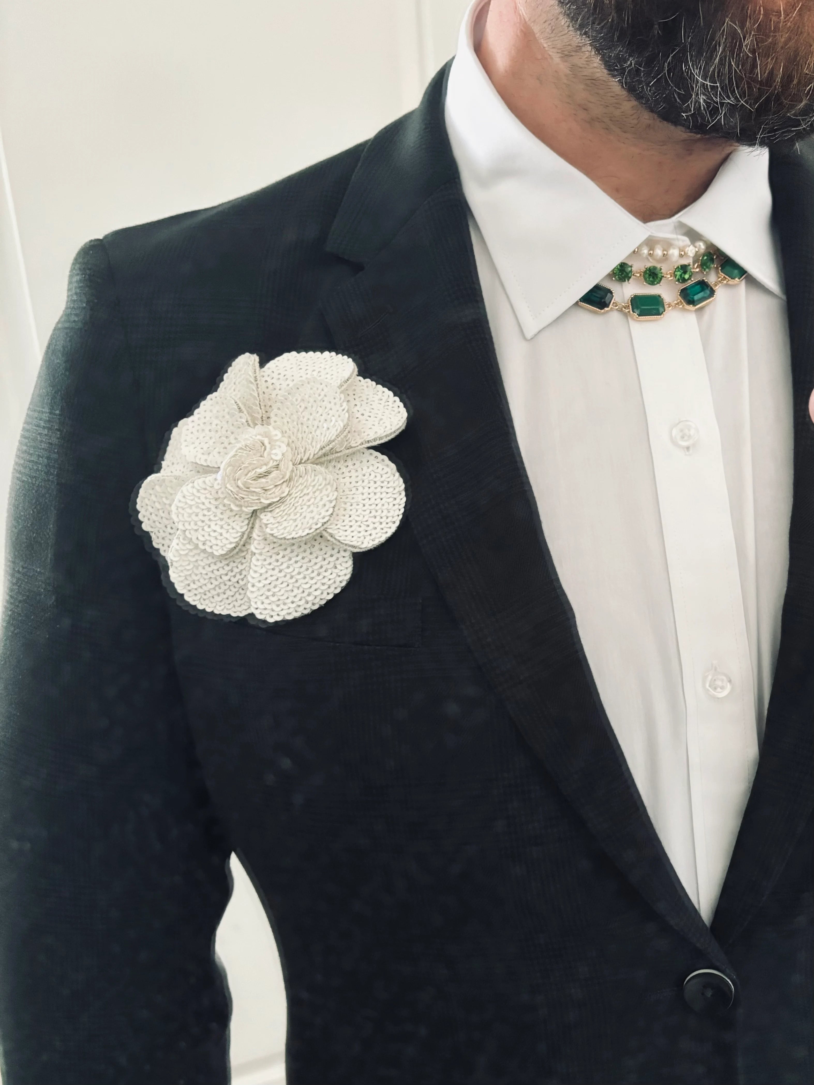 A close-up of a man wearing a black textured suit jacket, white dress shirt, and a decorative green and gold necktie, featuring a large white Broochella Scintillante sequin  flower brooch on the chest.