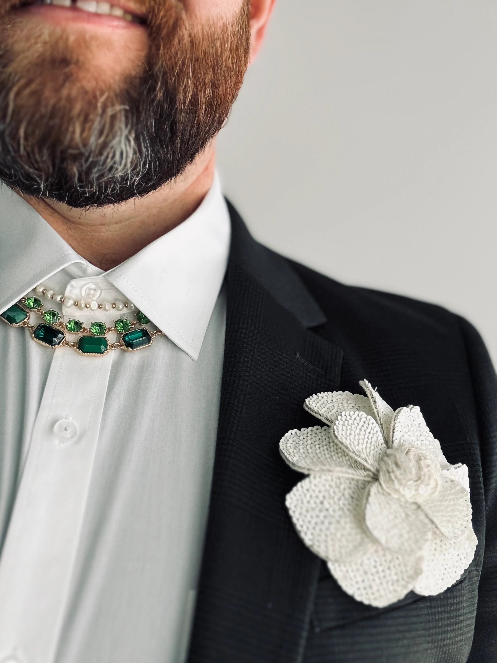 A close-up of a man wearing a black textured suit jacket, white dress shirt, and a decorative green and gold necktie, featuring a large white Broochella Scintillante sequin  flower brooch on the chest.