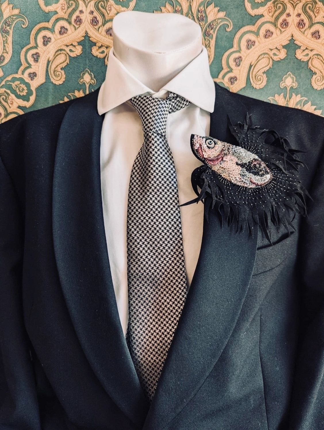A black suit jacket displayed on a mannequin torso with a white dress shirt, patterned grey tie, and a decorative Broochella Men's siamese black feathered fish-shaped lapel pin against ornate green and gold damask wallpaper.