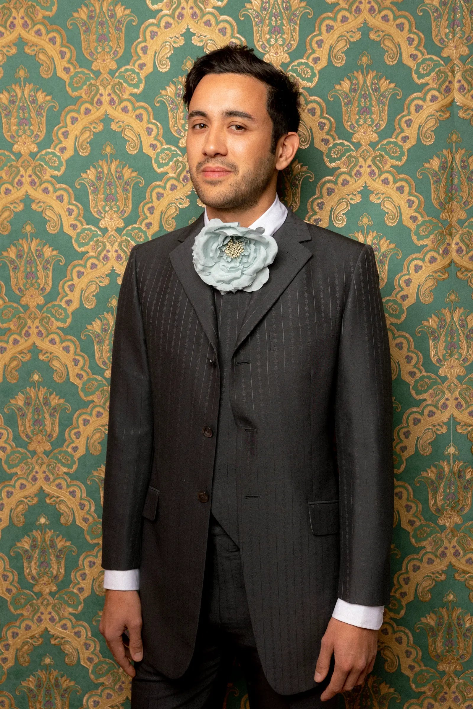 A man wearing a dark grey pinstripe suit, white dress shirt, and a Broochella signorina fioritina large, pale blue fabric flower brooch, standing in front of a green and gold damask patterned wallpaper background.
