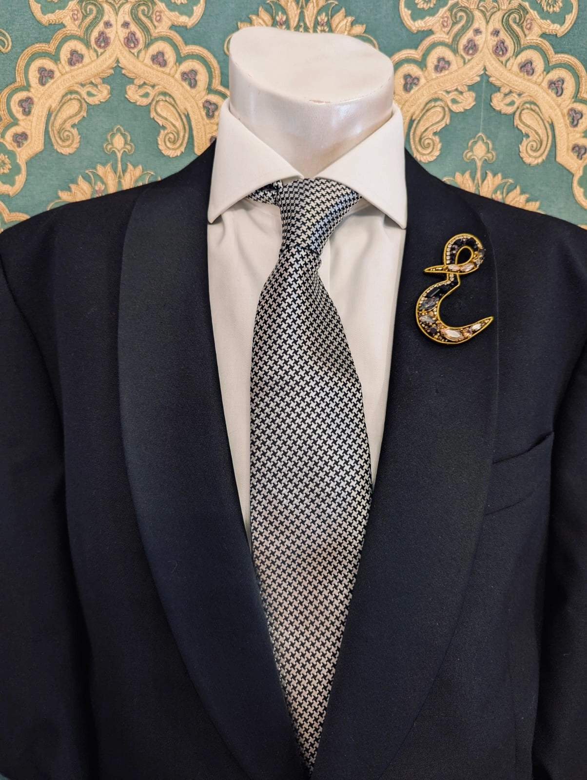 Broochella men's hand beaded E initial brooch in black and gold beads and sequins. Pinned to a men's mannequin wearing a black suit and a tie. The background is green and gold damask wallpaper.