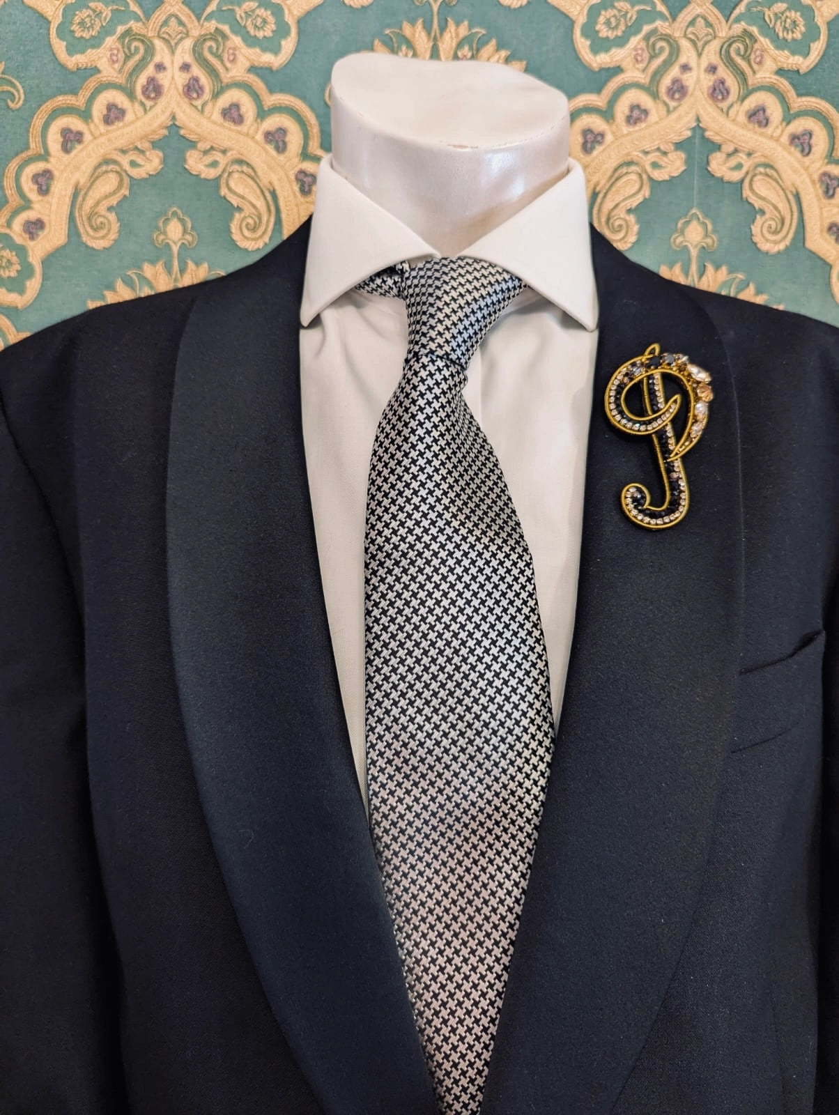 Broochella men's hand beaded P initial brooch in black and gold beads and sequins. Pinned to a men's mannequin wearing a black suit and a tie. The background is green and gold damask wallpaper.