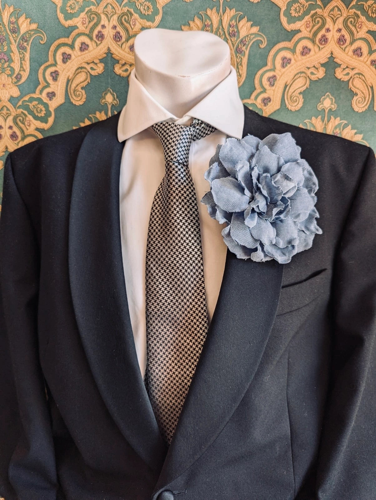A dark navy suit jacket displayed on a white mannequin torso, styled with a white dress shirt, a grey patterned tie, and a Broochella men's sky blue denim handmade flower brooch, set against a teal and gold damask wallpaper background.