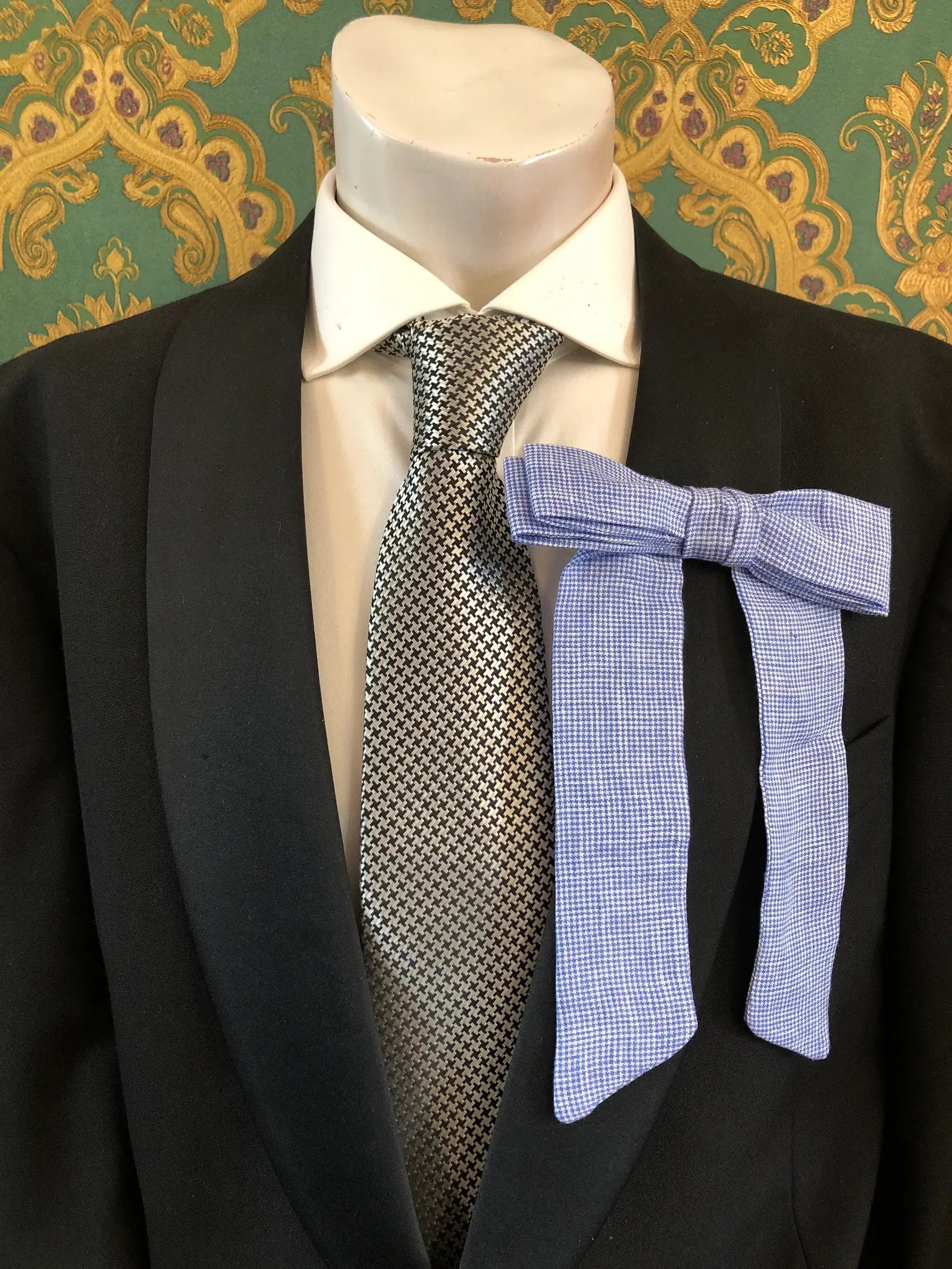 A Broochella Men's handmade Sofia Linen bow brooch in blue check styled on a mannequin torso wearing a black suit jacket, white dress shirt, and a black-and-white checkered tie. The background is green and gold damask wallpaper. 