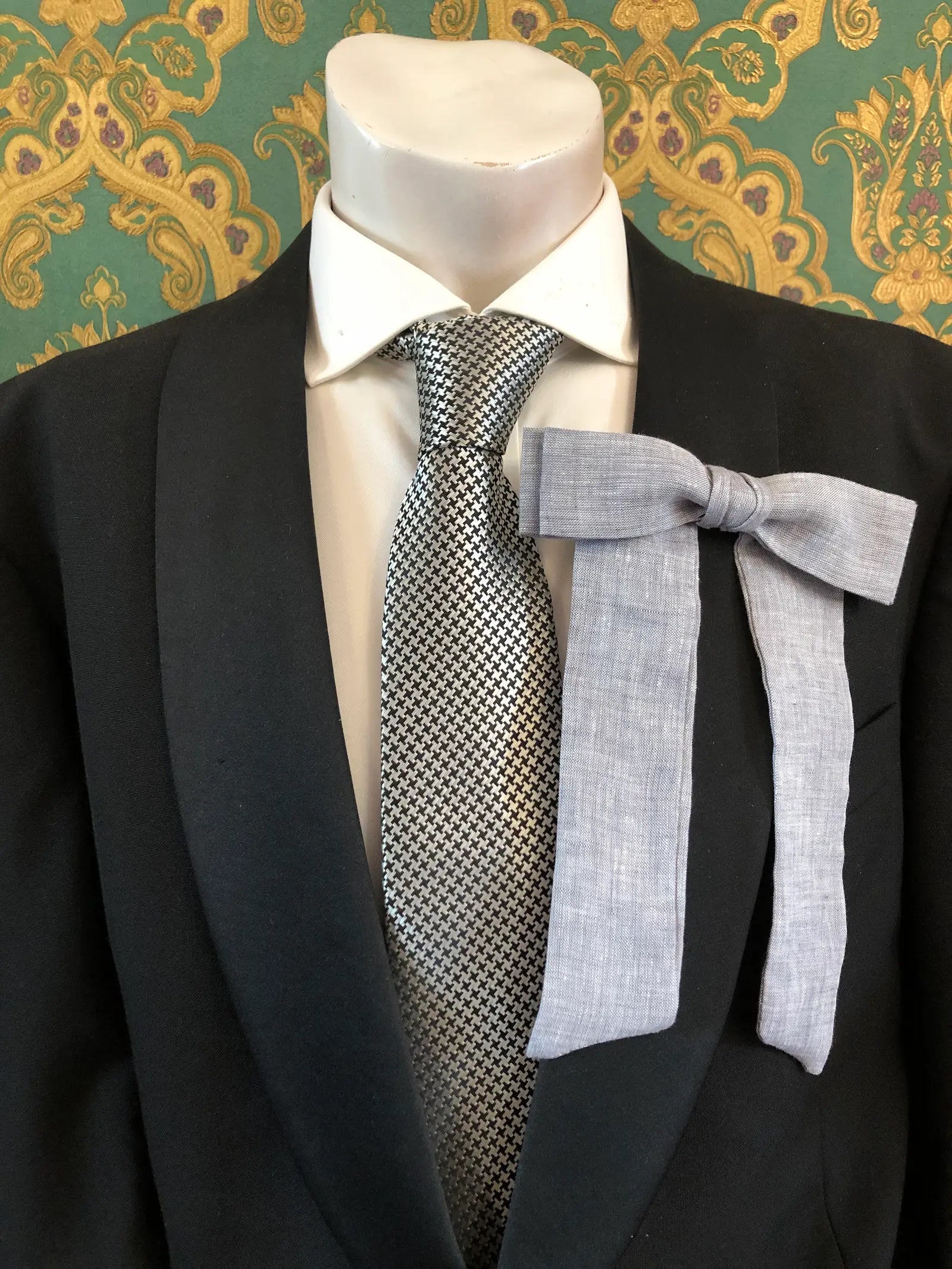 A Broochella Men's handmade Sofia Linen bow brooch in grey styled on a mannequin torso wearing a black suit jacket, white dress shirt, and a black-and-white checkered tie. The background is green and gold damask wallpaper. 