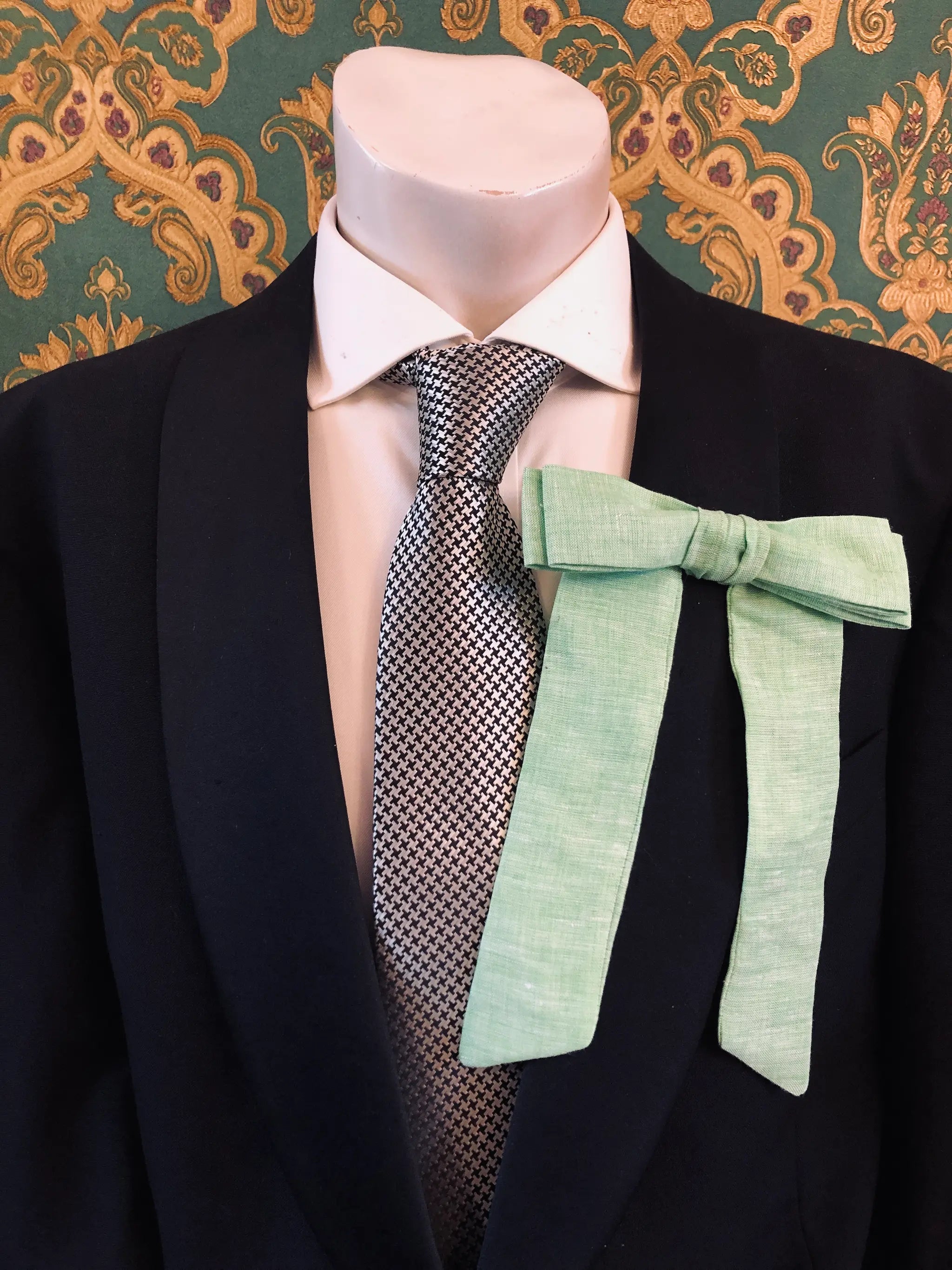 A Broochella Men's handmade Sofia Linen bow brooch in lime green styled on a mannequin torso wearing a black suit jacket, white dress shirt, and a black-and-white checkered tie. The background is green and gold damask wallpaper. 