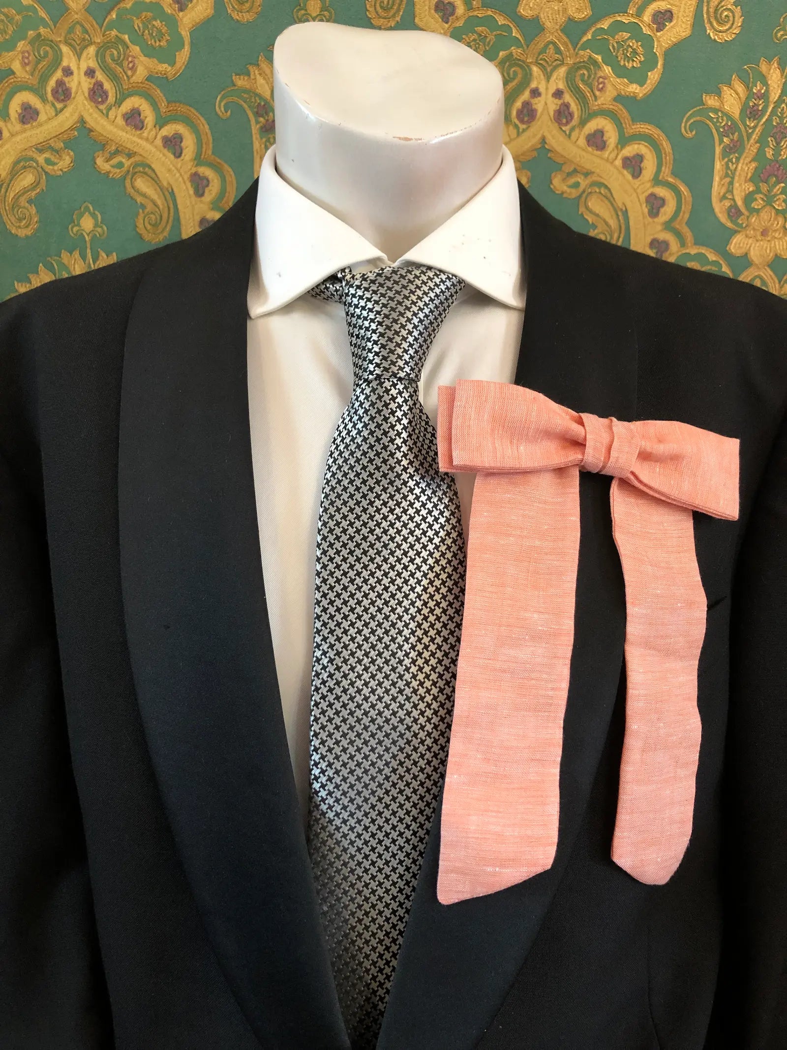 A Broochella Men's handmade Sofia Linen bow brooch in peach styled on a mannequin torso wearing a black suit jacket, white dress shirt, and a black-and-white checkered tie. The background is green and gold damask wallpaper. 