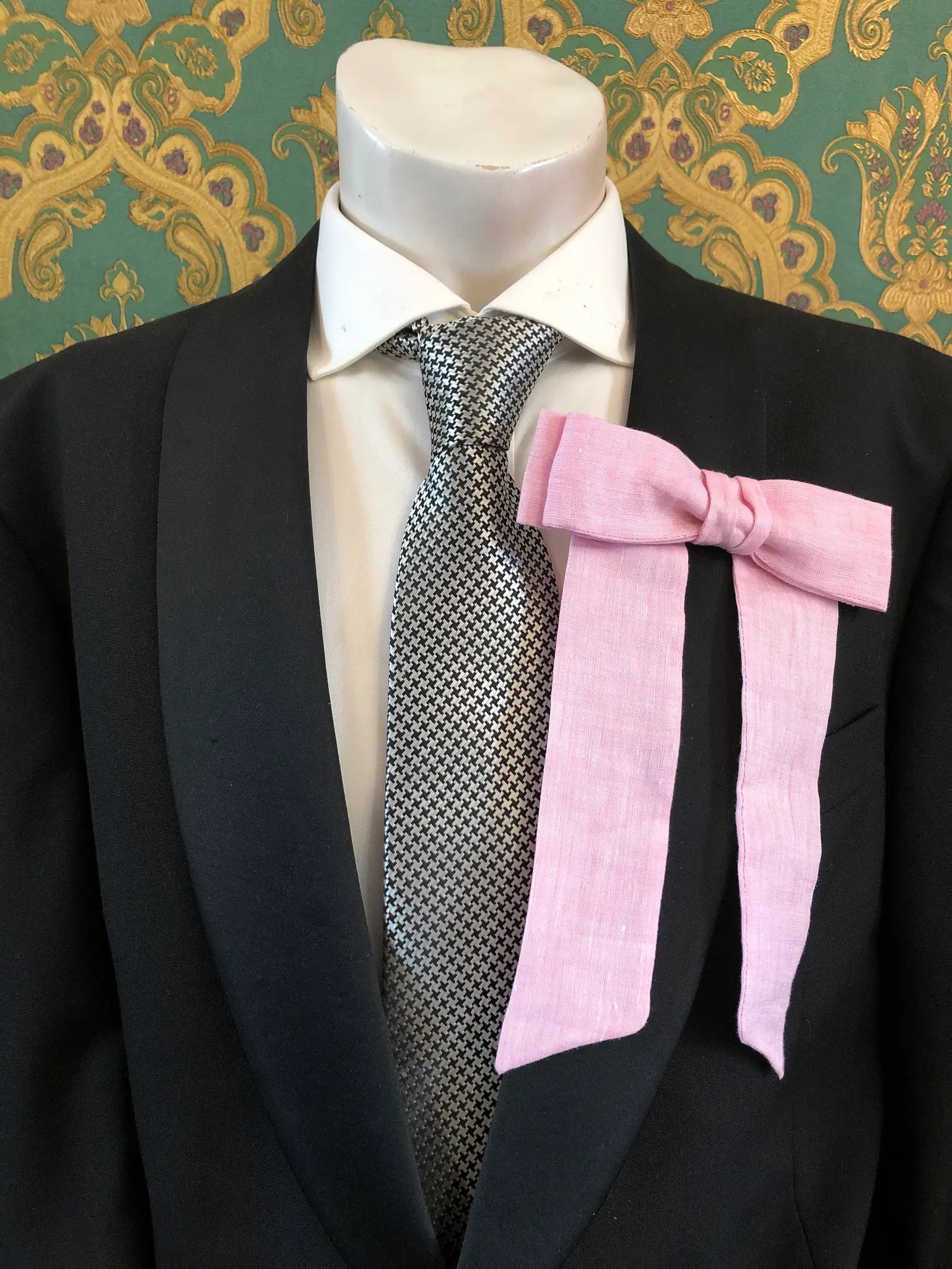 A Broochella Men's handmade Sofia Linen bow brooch in pink styled on a mannequin torso wearing a black suit jacket, white dress shirt, and a black-and-white checkered tie. The background is green and gold damask wallpaper. 
