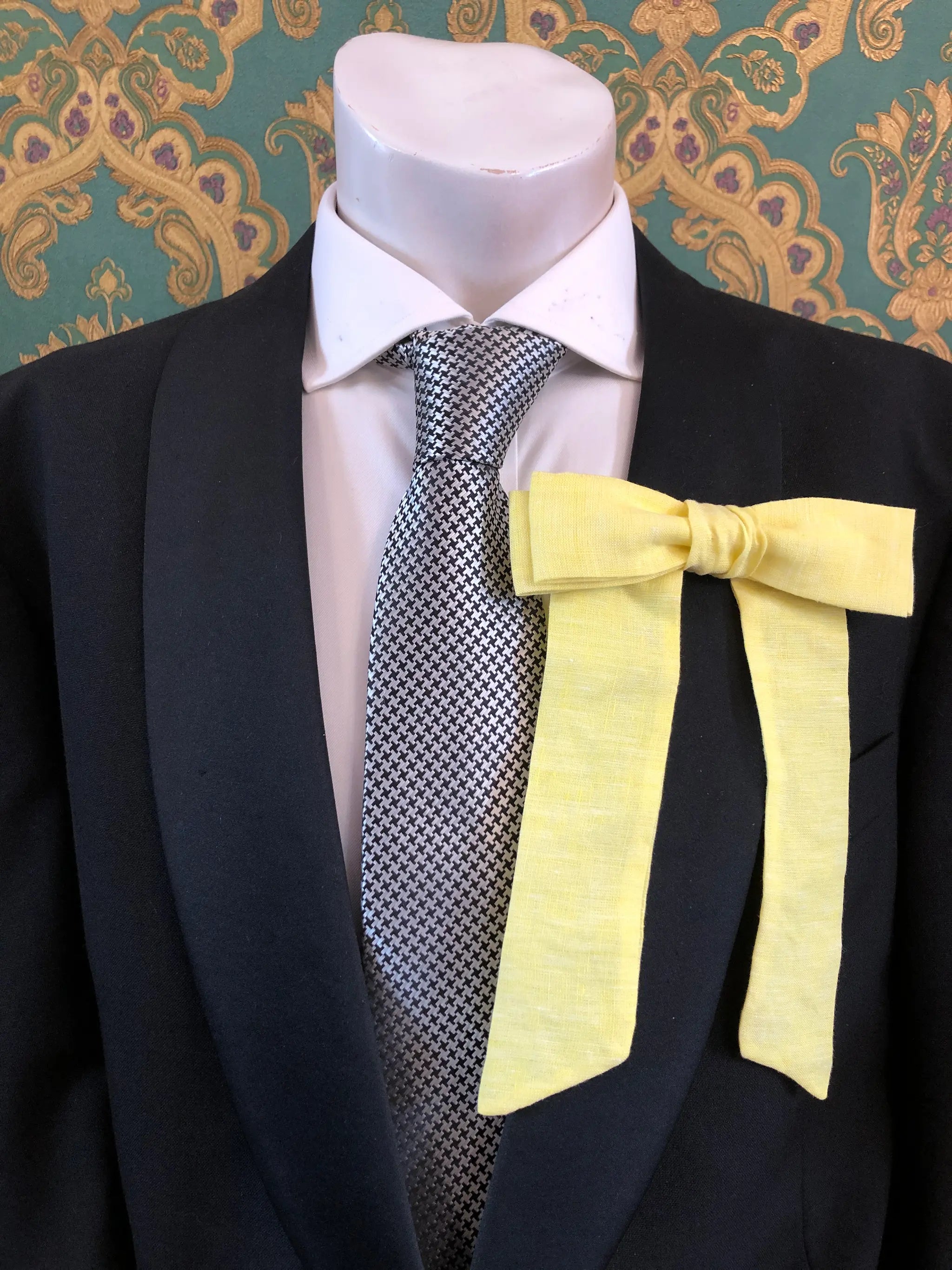 A Broochella Men's handmade Sofia Linen bow brooch in yellow styled on a mannequin torso wearing a black suit jacket, white dress shirt, and a black-and-white checkered tie. The background is green and gold damask wallpaper. 