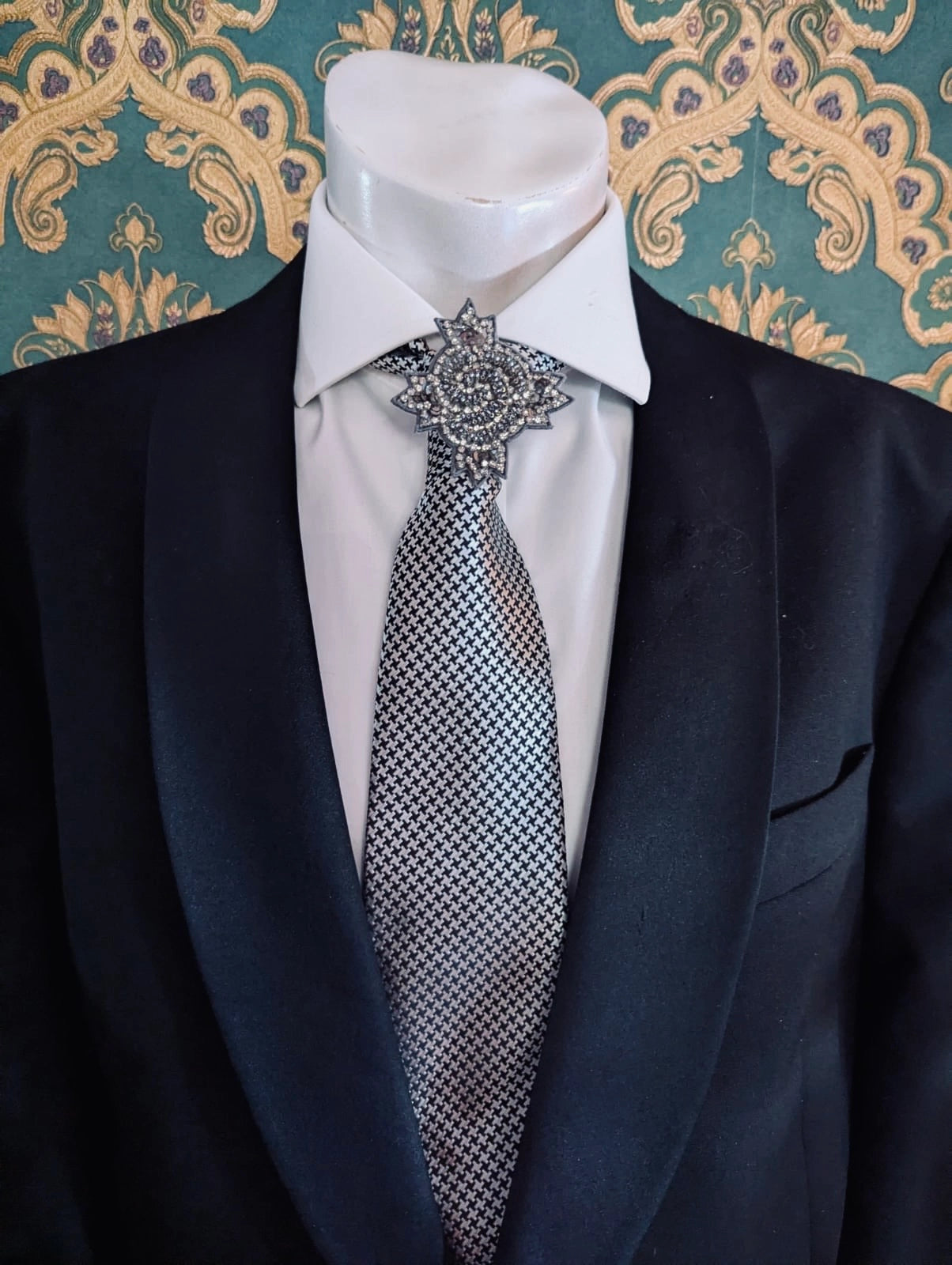 Broochella men's handmade, hand beaded designer, star bust brooch in beads and sequins. Pinned on a men's mannequin wearing a black suit and a houndstooth tie. The background is green and gold damask wallpaper. 