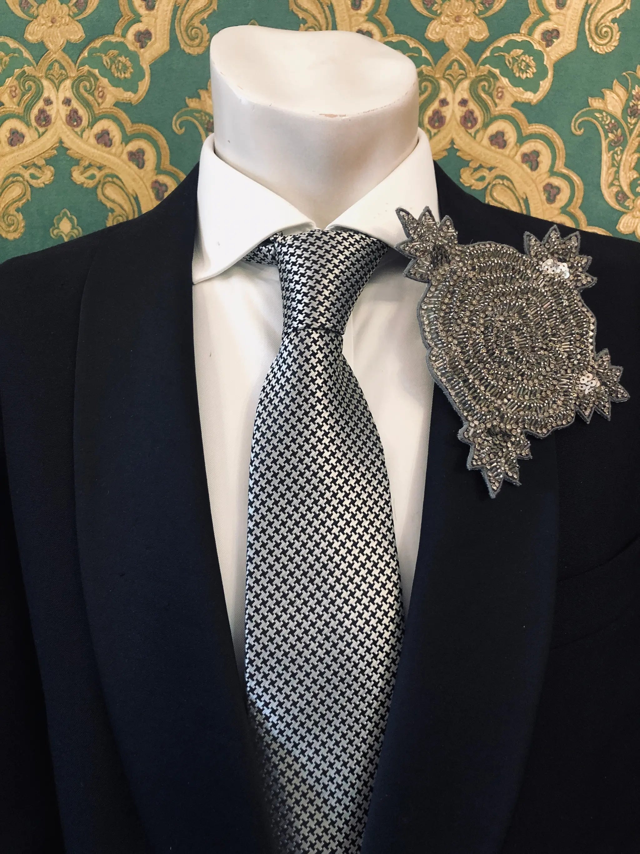 A close-up of a mannequin torso wearing a black suit jacket, white dress shirt, and a silver-and-black patterned tie, with a large Broochella Stella hand beaded ornate silver brooch pinned to the lapel. The background is patterned green and gold wallpaper.