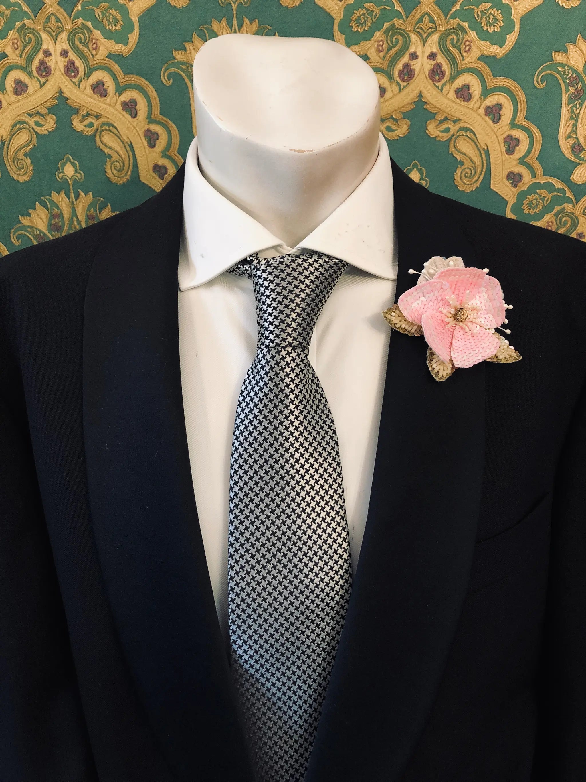 A Broochella Men's Tesorino Ingioiellato pink fabric flower brooch with gold metallic accents, styled on a white dress shirt and patterned gray tie inside a dark navy suit jacket, displayed on a mannequin torso against a green and gold patterned wallpaper background.