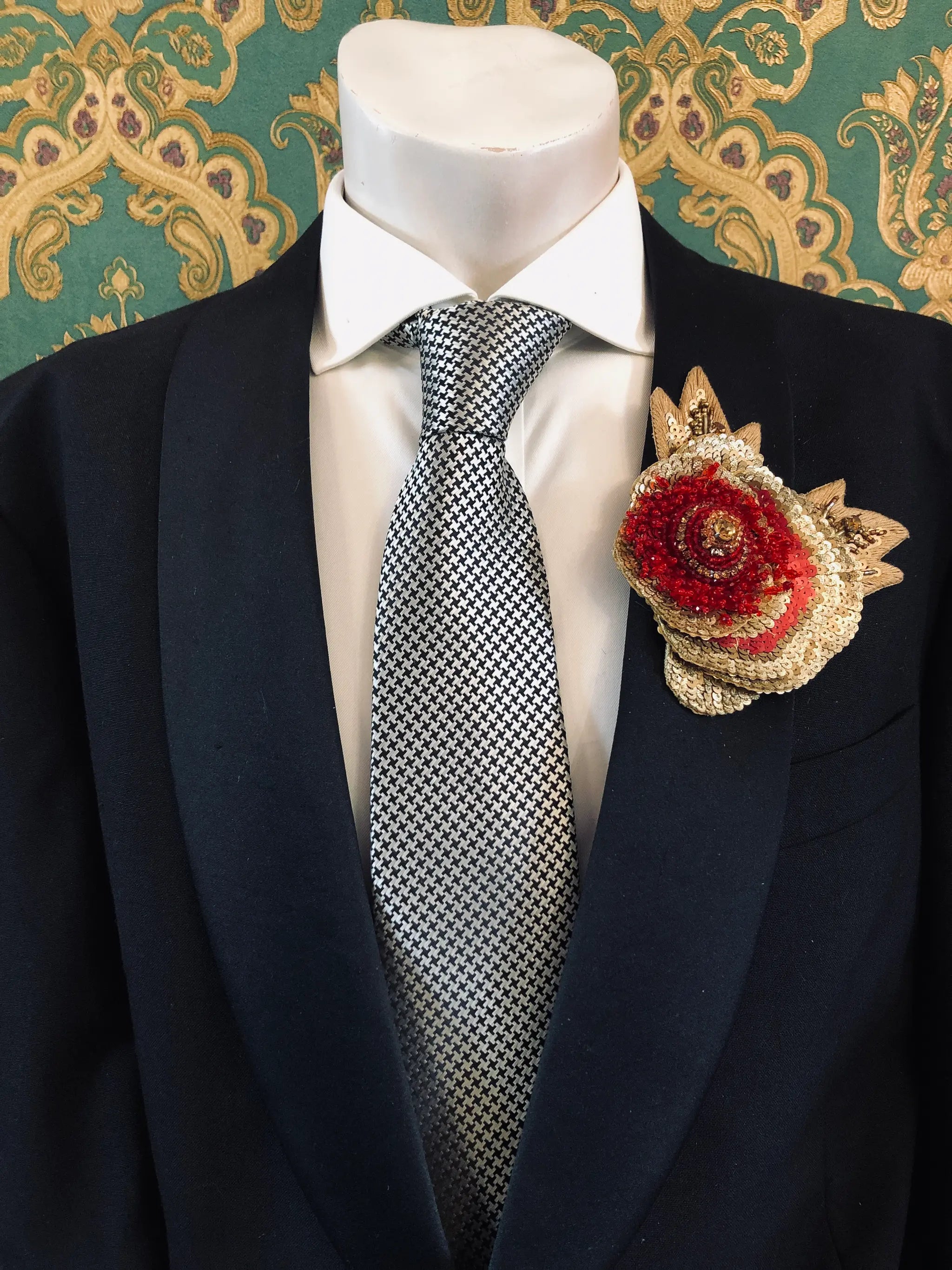 A formal men's suit ensemble displayed on a mannequin torso, featuring a black suit jacket, a white dress shirt, a silver and black checkered tie, and a Broochella Men's Tonina Tesorina red and gold hand beaded flower brooch