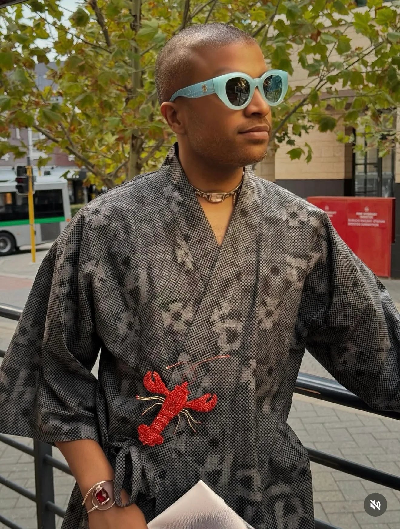 A man wearing a gray and black patterned kimono-style robe with a Broochella hand beaded Tropicana red lobster brooch at the waist, light blue sunglasses, and a gold chain necklace, standing outdoors on a city street.