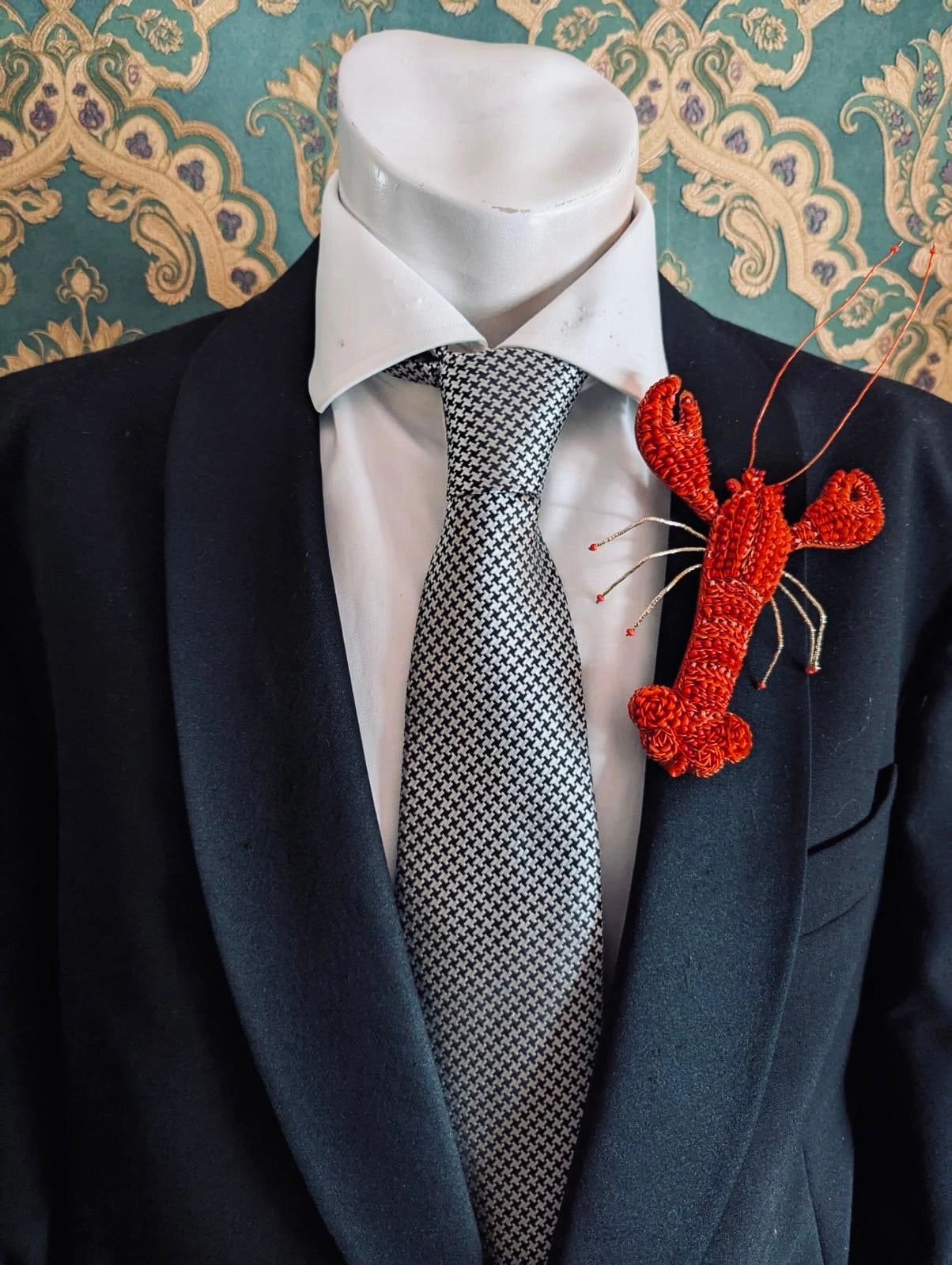 A Broochella Tropicana red hand beaded lobster brooch displayed on a white shirt and patterned grey tie, worn on a black suit jacket mannequin torso against a teal damask patterned background.