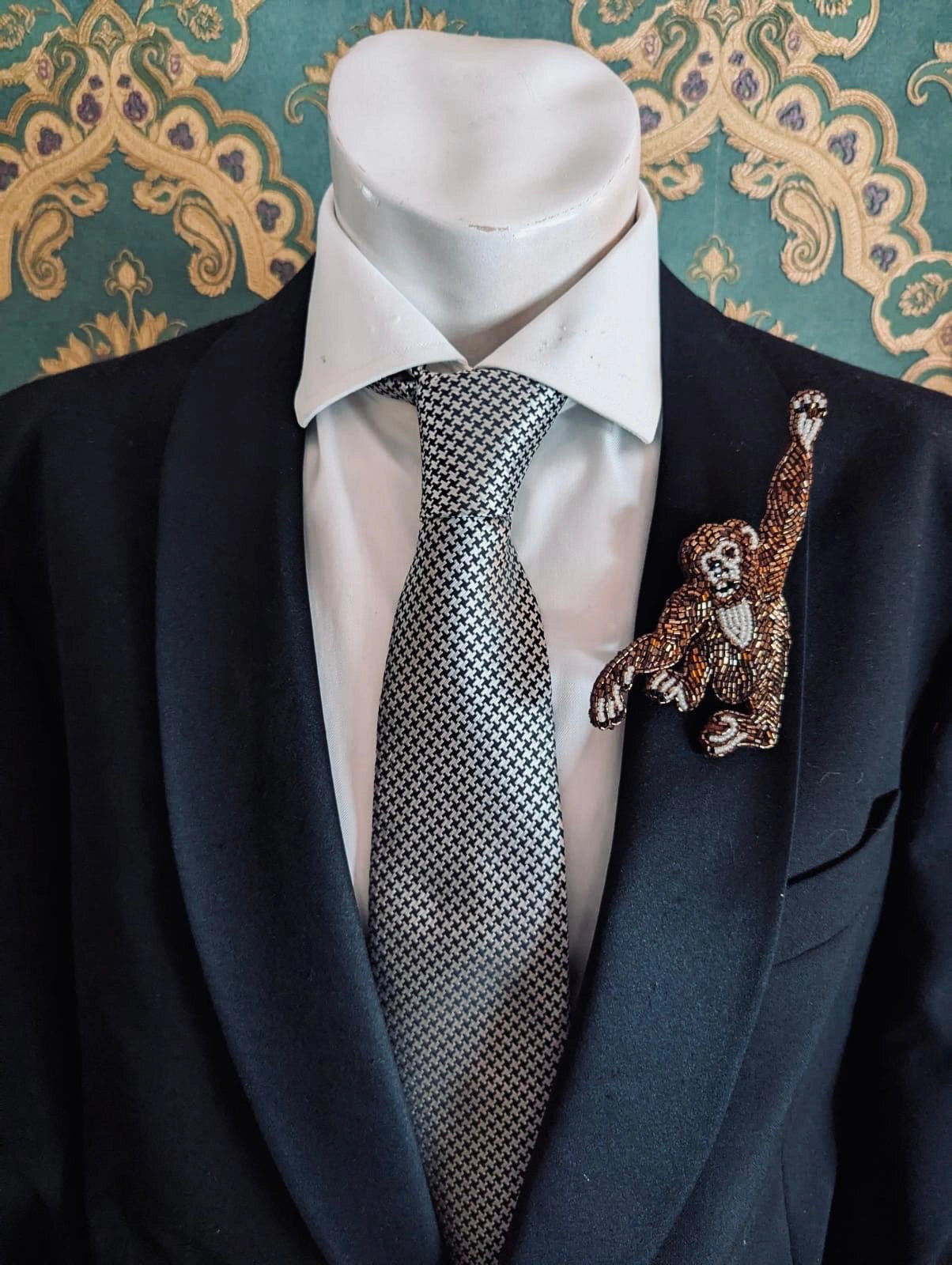 Black suit with white shirt, patterned tie, and Broochella men's Tropicana collection hand beaded monkey brooch against a decorative wall.