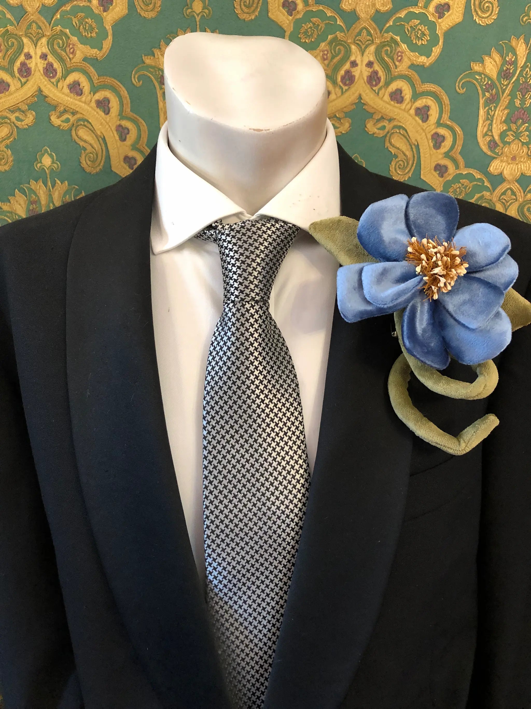 A close-up of a mannequin torso wearing a black suit jacket, white dress shirt, and a black-and-white patterned tie, with a large Broochella Men's Velluto  blue velvet flower brooch pinned to the left lapel. The background is a green and gold damask wallpaper.
