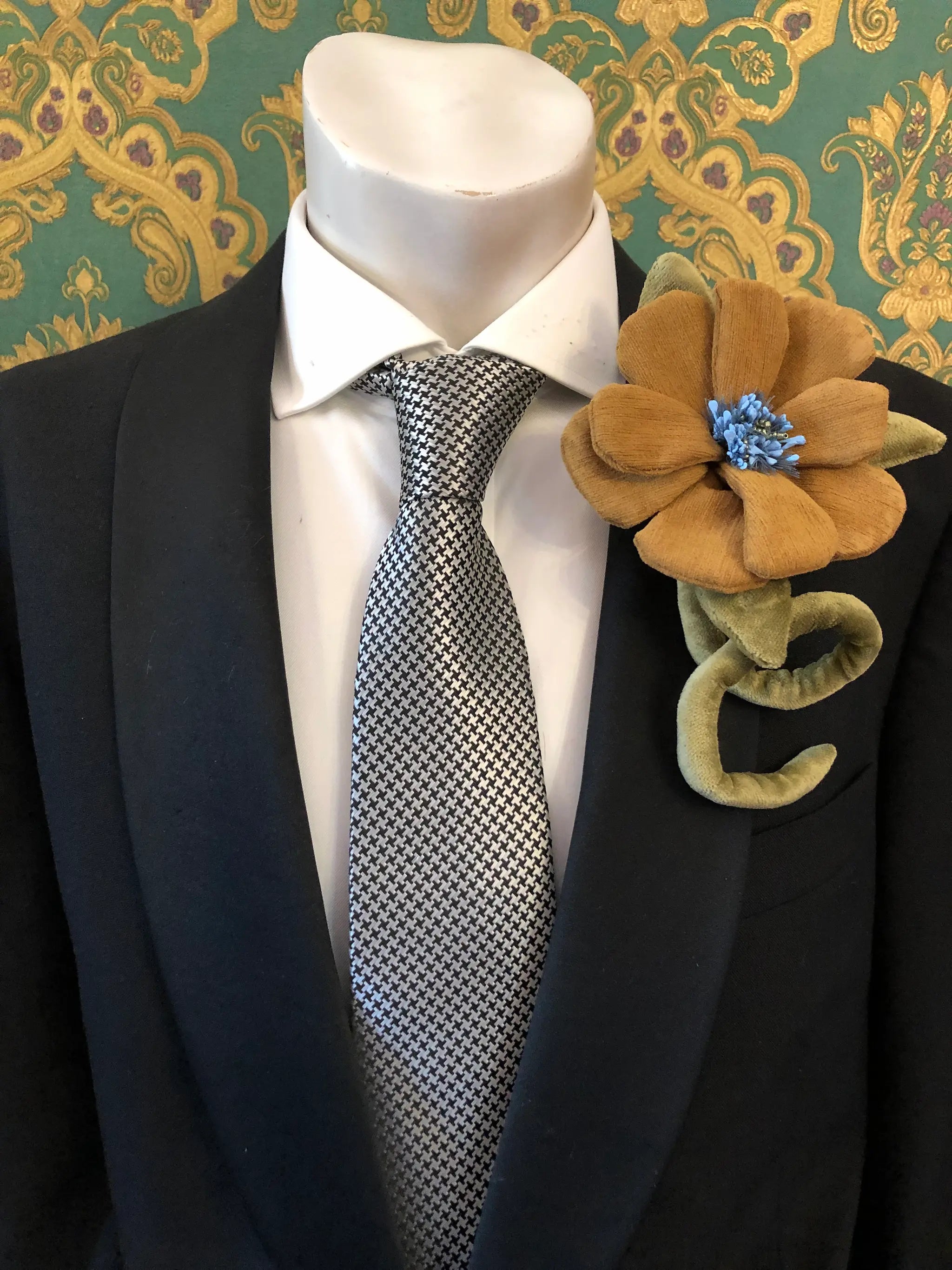 A close-up of a mannequin torso wearing a black suit jacket, white dress shirt, and a black-and-white patterned tie, with a large Broochella Men's Velluto brown velvet flower brooch pinned to the left lapel. The background is a green and gold damask wallpaper.