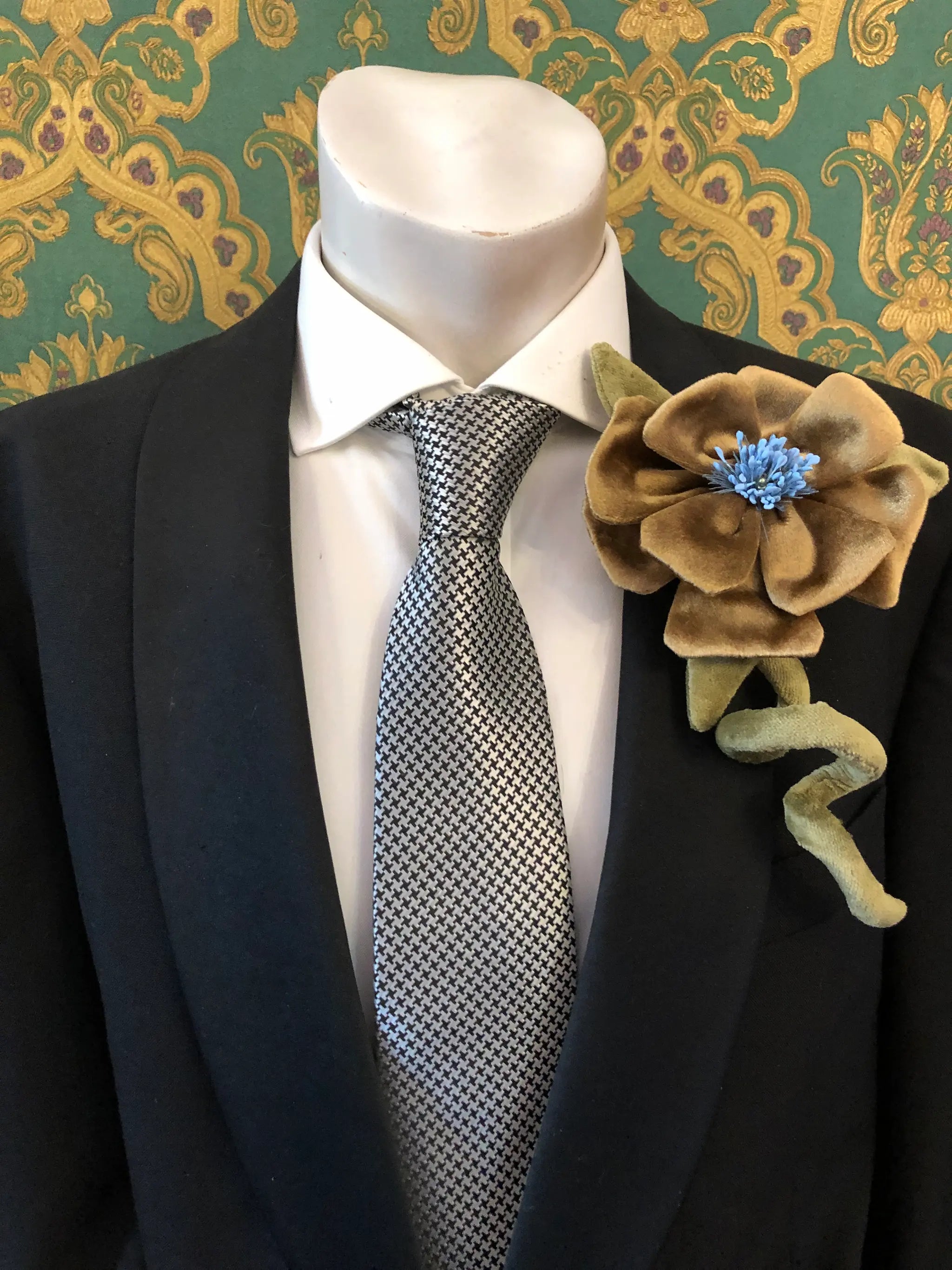 A close-up of a mannequin torso wearing a black suit jacket, white dress shirt, and a black-and-white patterned tie, with a large Broochella Men's Velluto gold velvet flower brooch pinned to the left lapel. The background is a green and gold damask wallpaper.
