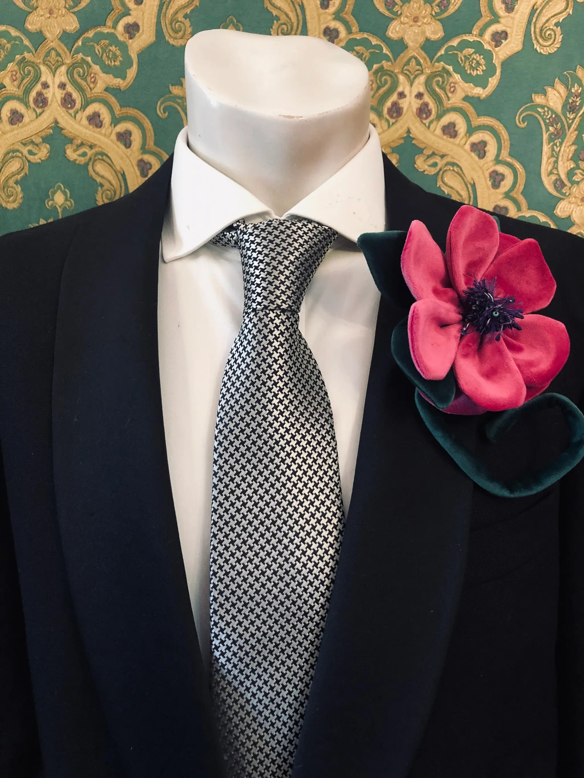 A close-up of a mannequin torso wearing a black suit jacket, white dress shirt, and a black-and-white patterned tie, with a large Broochella Men's Velluto  magenta velvet flower brooch pinned to the left lapel. The background is a green and gold damask wallpaper.