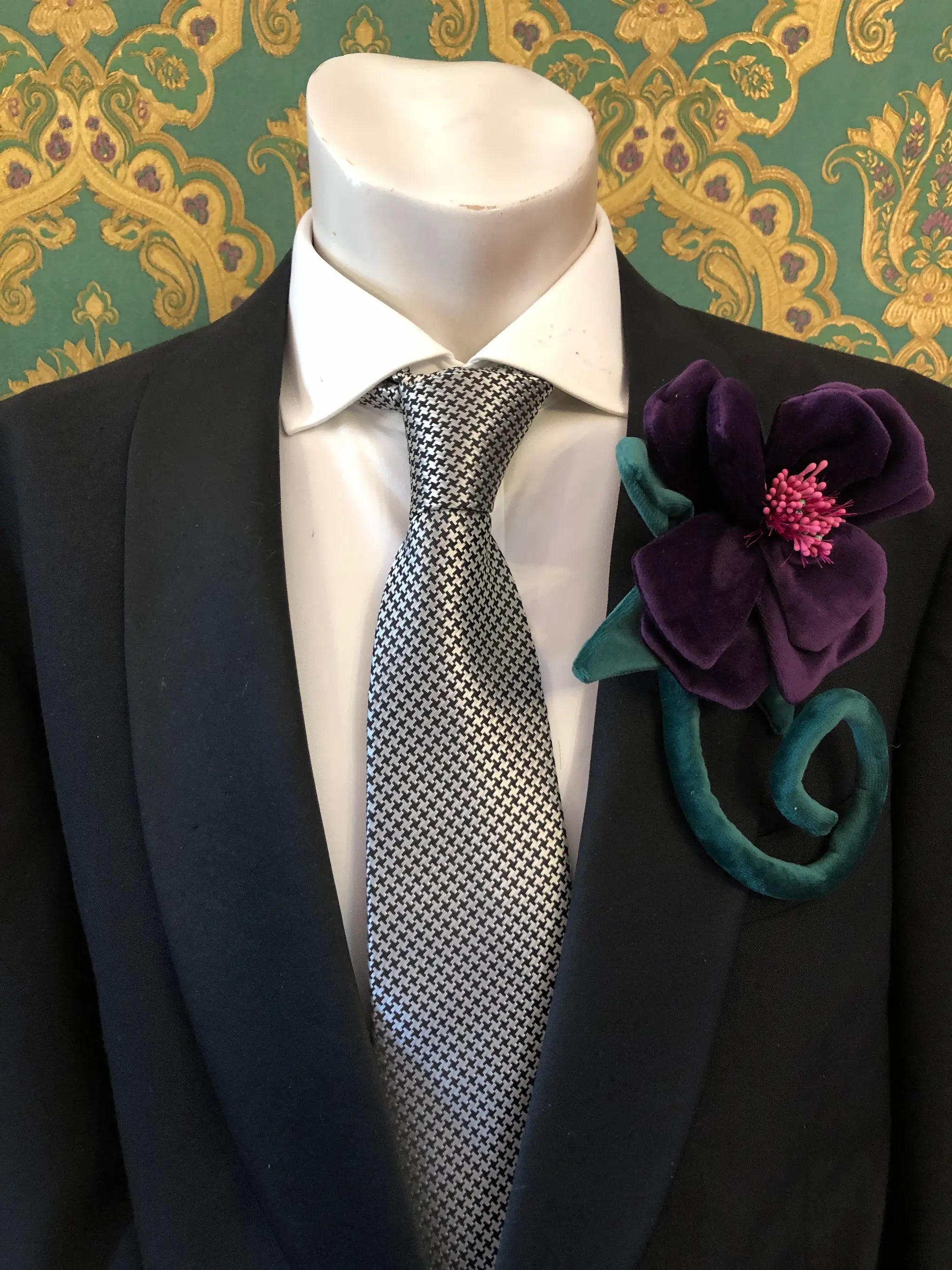 A close-up of a mannequin torso wearing a black suit jacket, white dress shirt, and a black-and-white patterned tie, with a large Broochella Men's Velluto purple velvet flower brooch pinned to the left lapel. The background is a green and gold damask wallpaper.