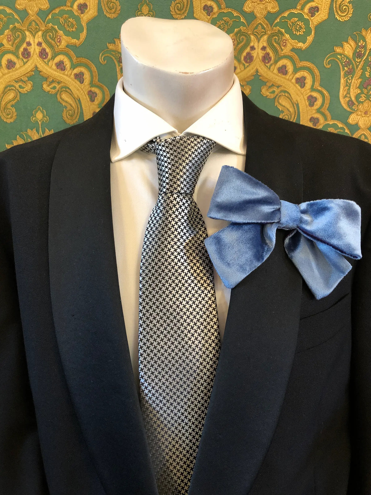 A Broochella Men's blue/purple velvet bow brooch displayed on a mannequin torso wearing a black suit jacket, white dress shirt, and a silver-and-black patterned tie. It is set against ornate green and gold patterned wallpaper.