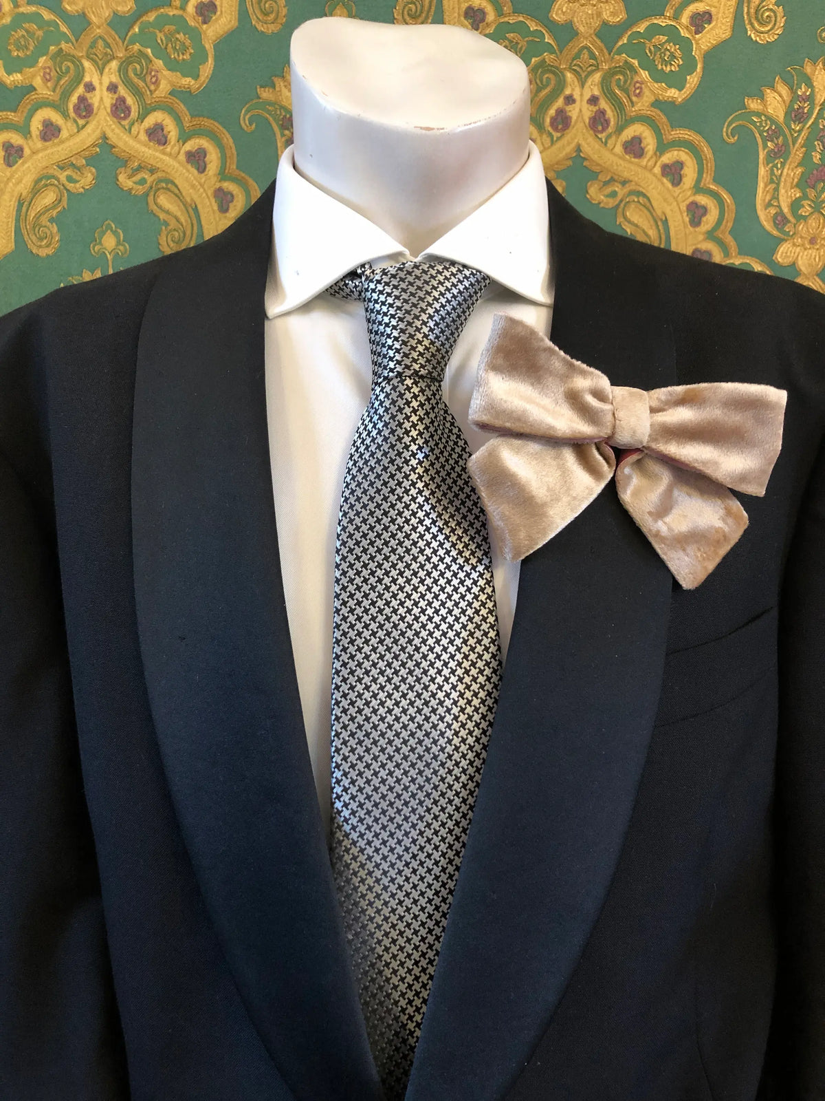 A Broochella Men's a champagne/pink velvet bow brooch displayed on a mannequin torso wearing a black suit jacket, white dress shirt, and a silver-and-black patterned tie. It is set against ornate green and gold patterned wallpaper.