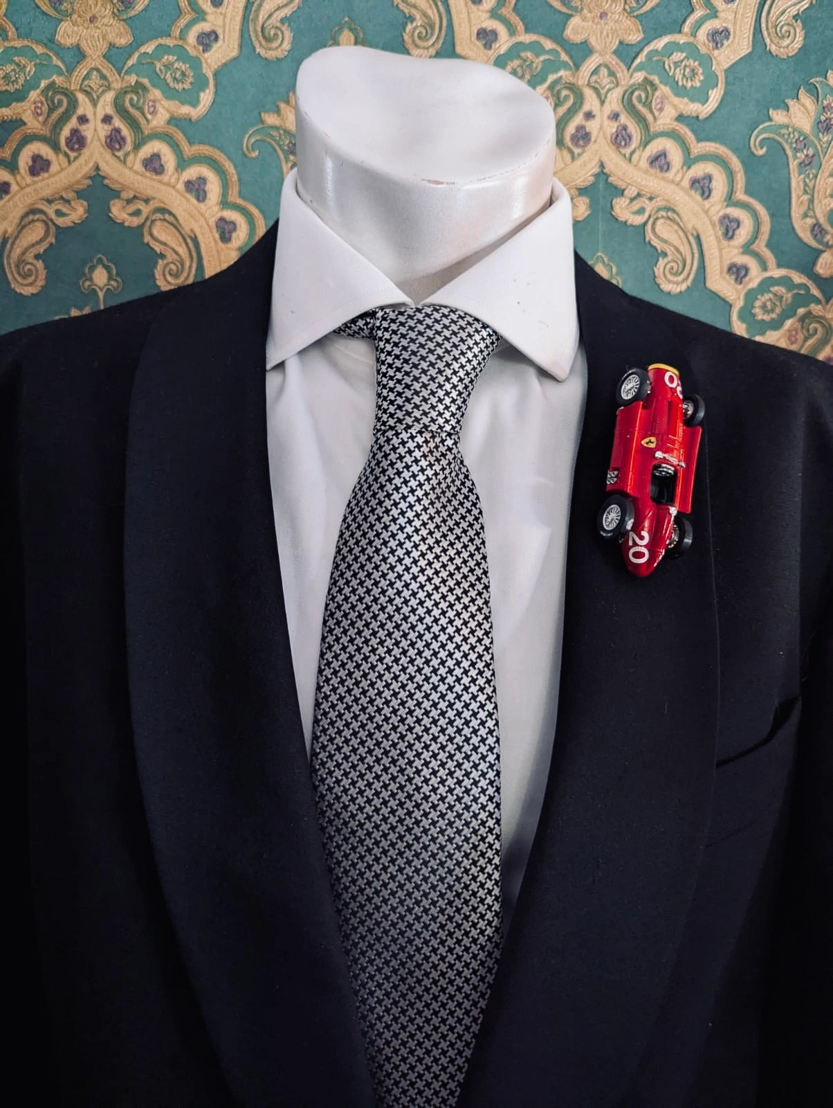 Broochella men's scuderia d'oro ferrari rosso classico 20 red  vintage formula one racing car brooch. Pinned on a men's mannequin wearing a black suit and a houndstooth tie. The background is green and gold damask wallpaper. 