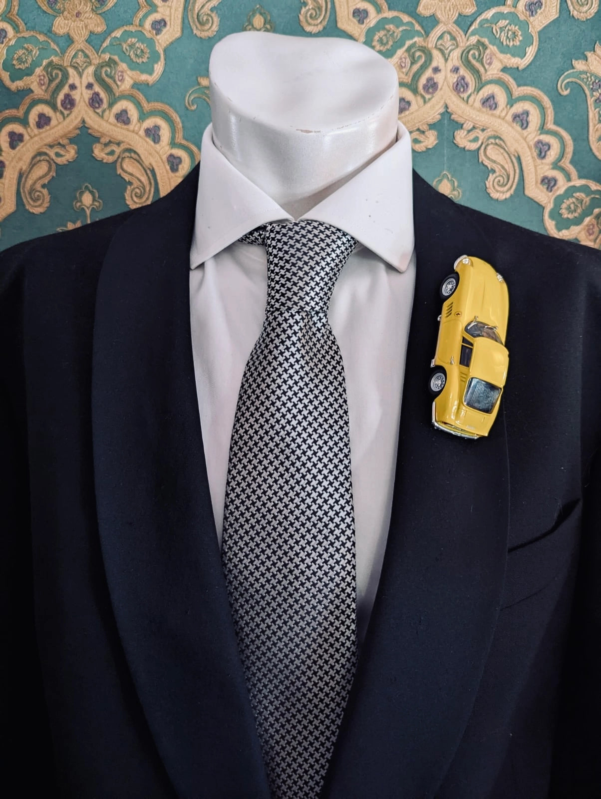 Broochella men's scuderia d'oro giallo fiorano number 275 vintage formula one racing car brooch. Pinned on a men's mannequin wearing a black suit and a houndstooth tie. The background is green and gold damask wallpaper. 