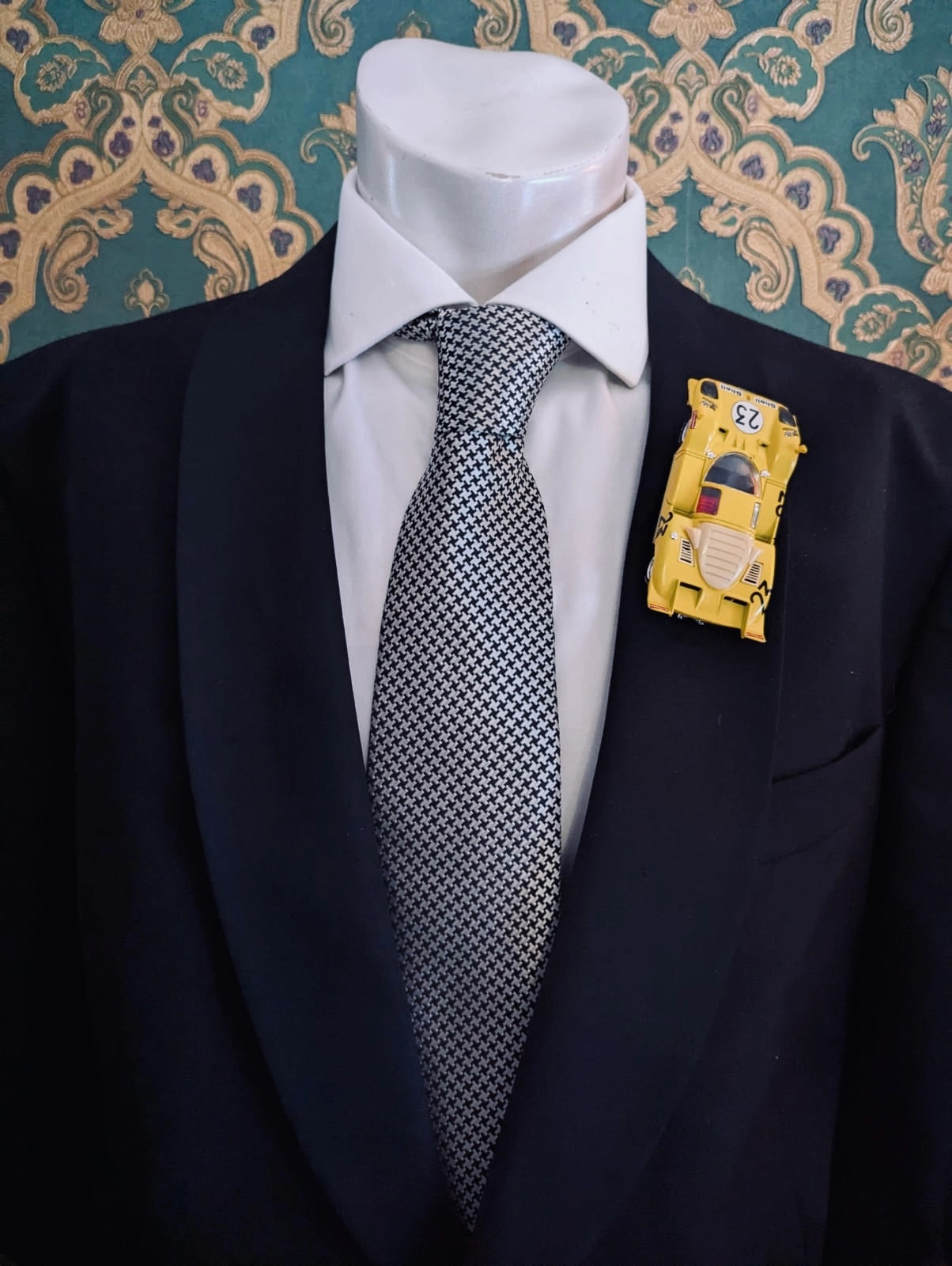 Broochella men's scuderia d'oro giallo sprint yellow number 23 vintage formula one racing car brooch. Pinned on a men's mannequin wearing a black suit and a houndstooth tie. The background is green and gold damask wallpaper. 