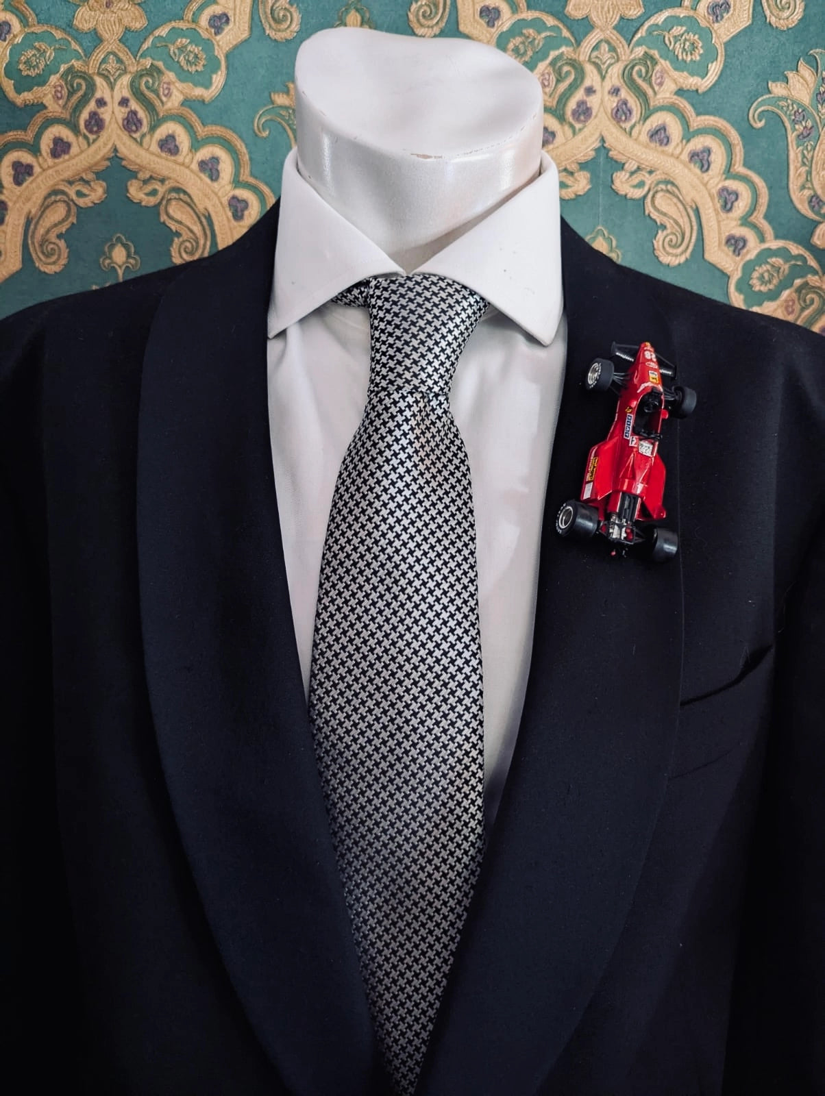 Broochella men's scuderia d'oro rosso monza 28 red, vintage formula one racing car brooch. Pinned on a men's mannequin wearing a black suit and a houndstooth tie. The background is green and gold damask wallpaper. 